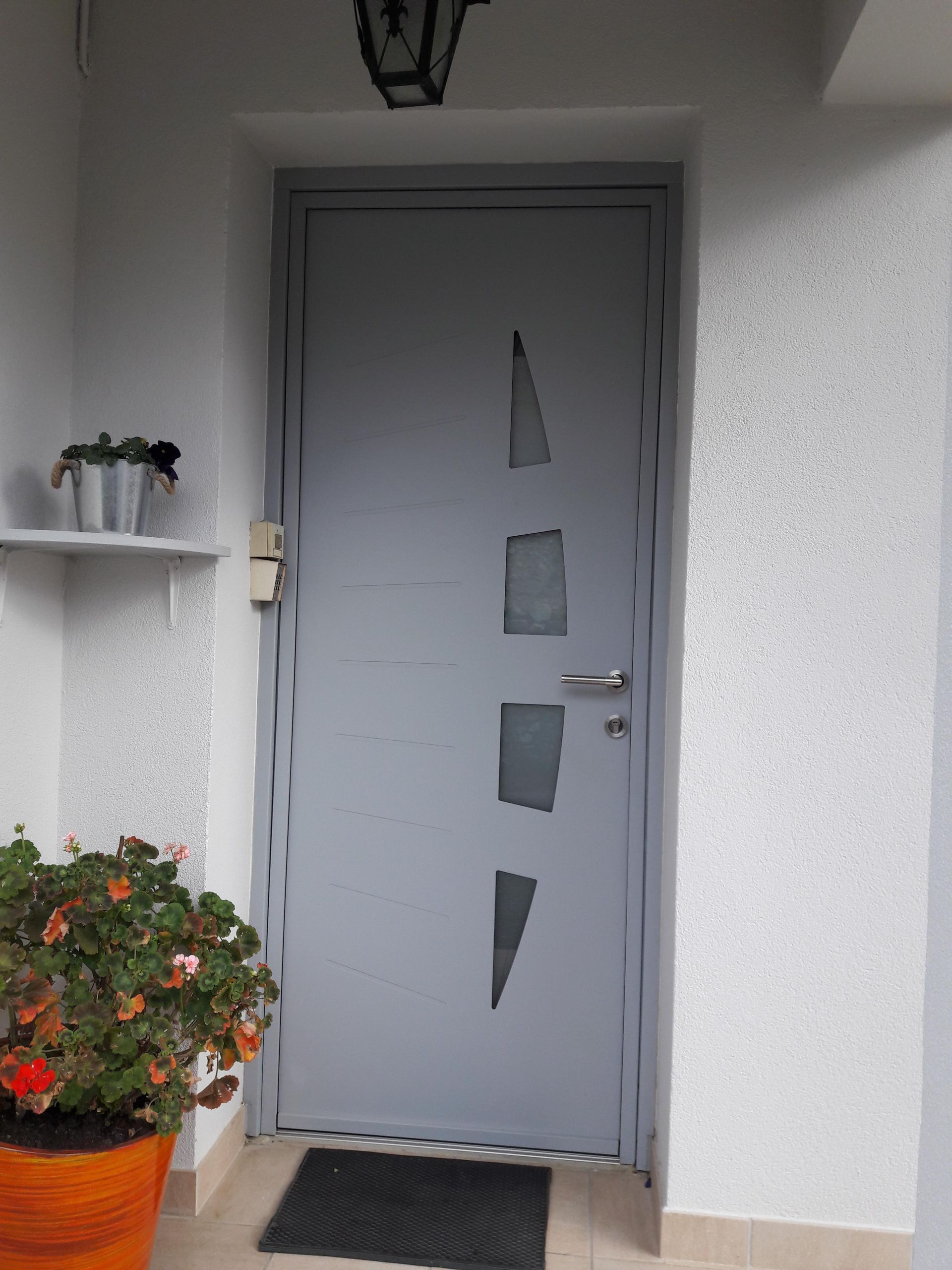 Une porte grise ornée de quatre découpes verticales irrégulières en verre, encastrée dans un mur extérieur blanc à côté d'une plante en pot.