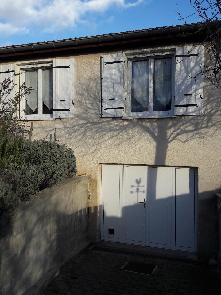 L'extérieur d'une maison beige avec des volets blancs à deux fenêtres, une porte de garage blanche et un arbre projetant son ombre sur le mur.