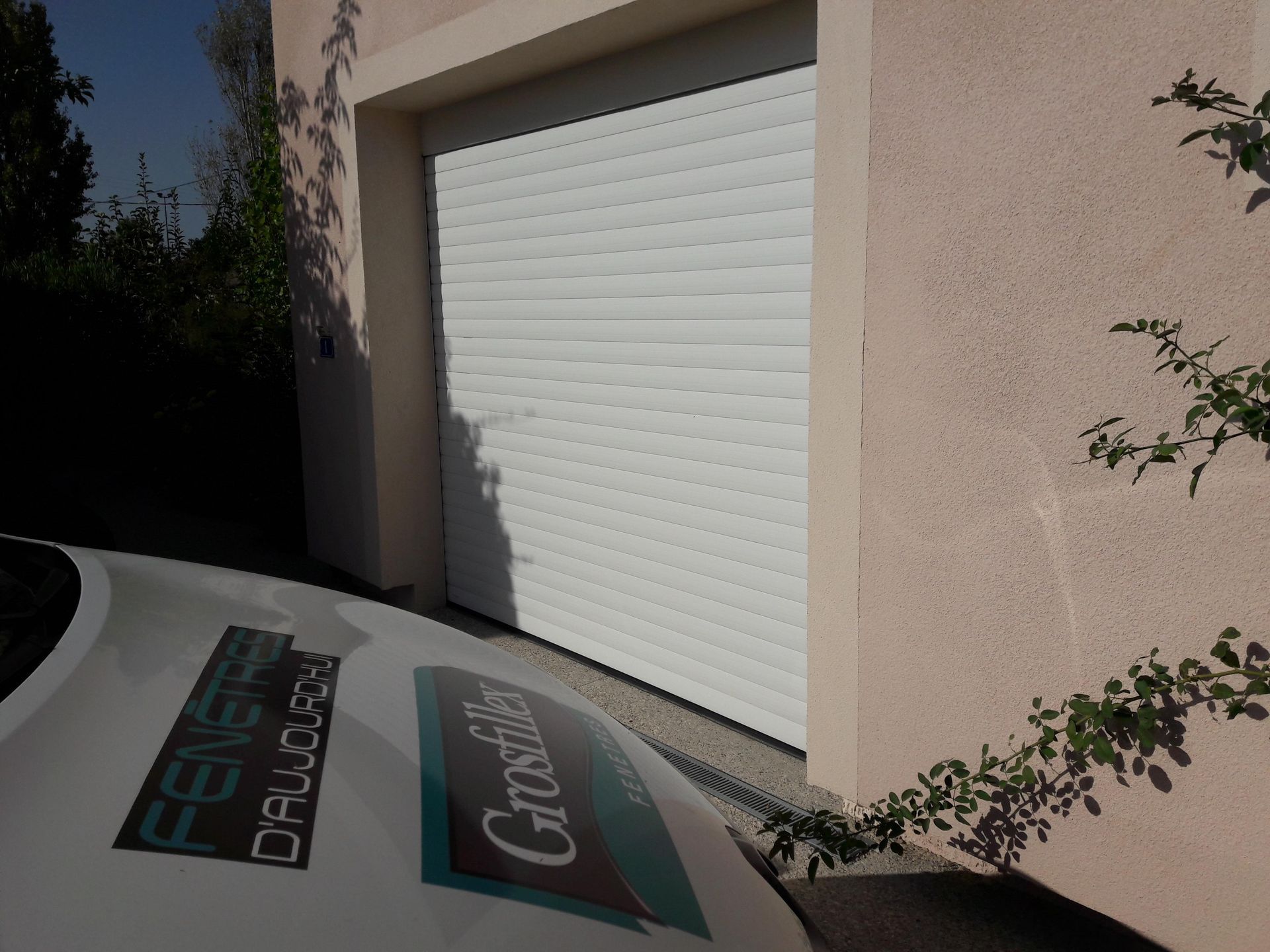 Porte de garage blanche fermée dans un bâtiment en stuc, avec le bord d'un véhicule de société aux couleurs de l'entreprise au premier plan.