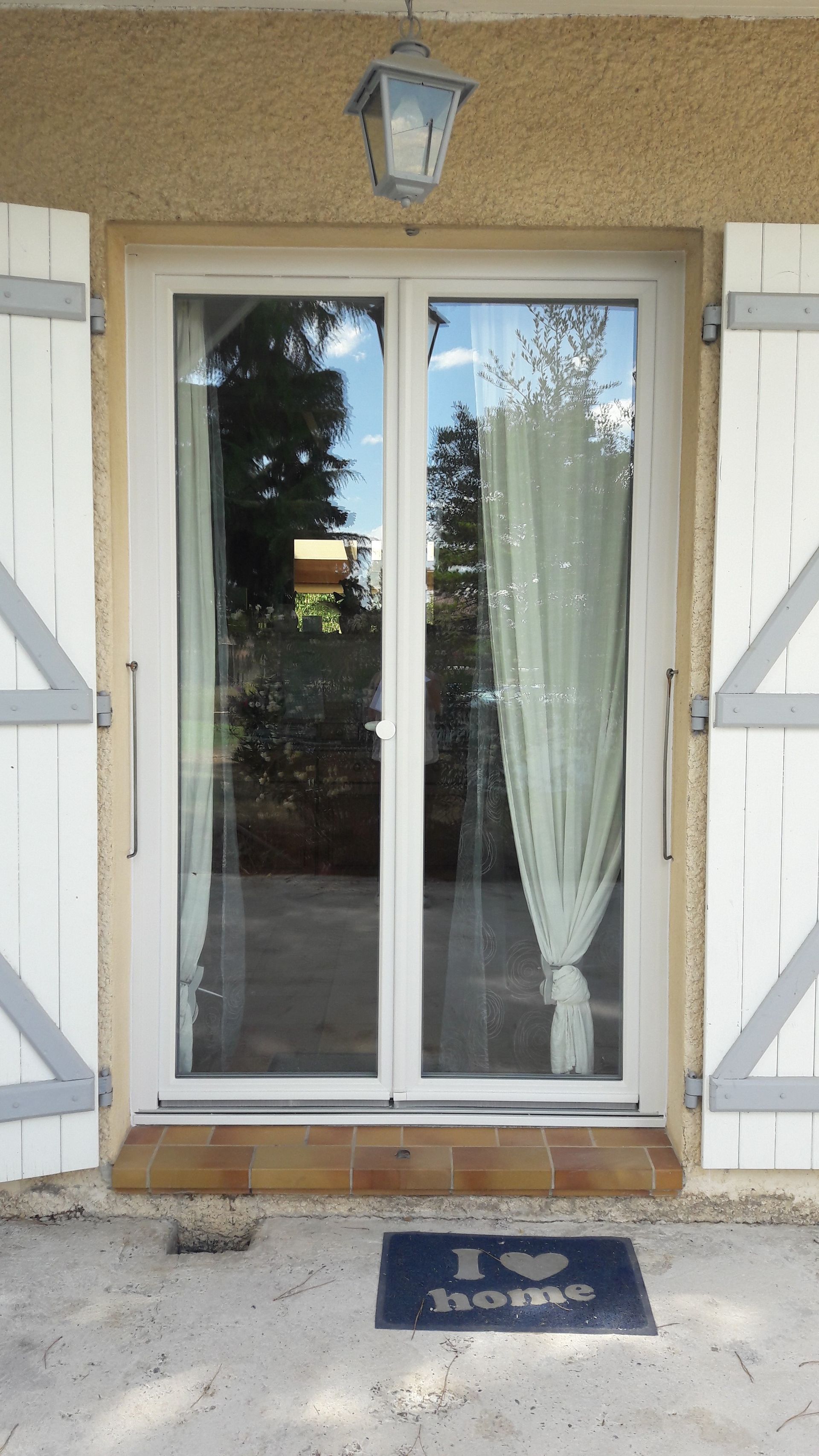 Portes-fenêtres blanches à volets blancs sur une façade beige, avec une lanterne au-dessus et un paillasson.