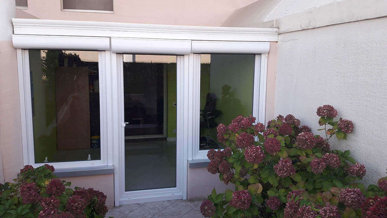 Une porte vitrée et deux fenêtres latérales à cadres blancs, partiellement recouvertes de volets, à côté d'un buisson d'hortensias.