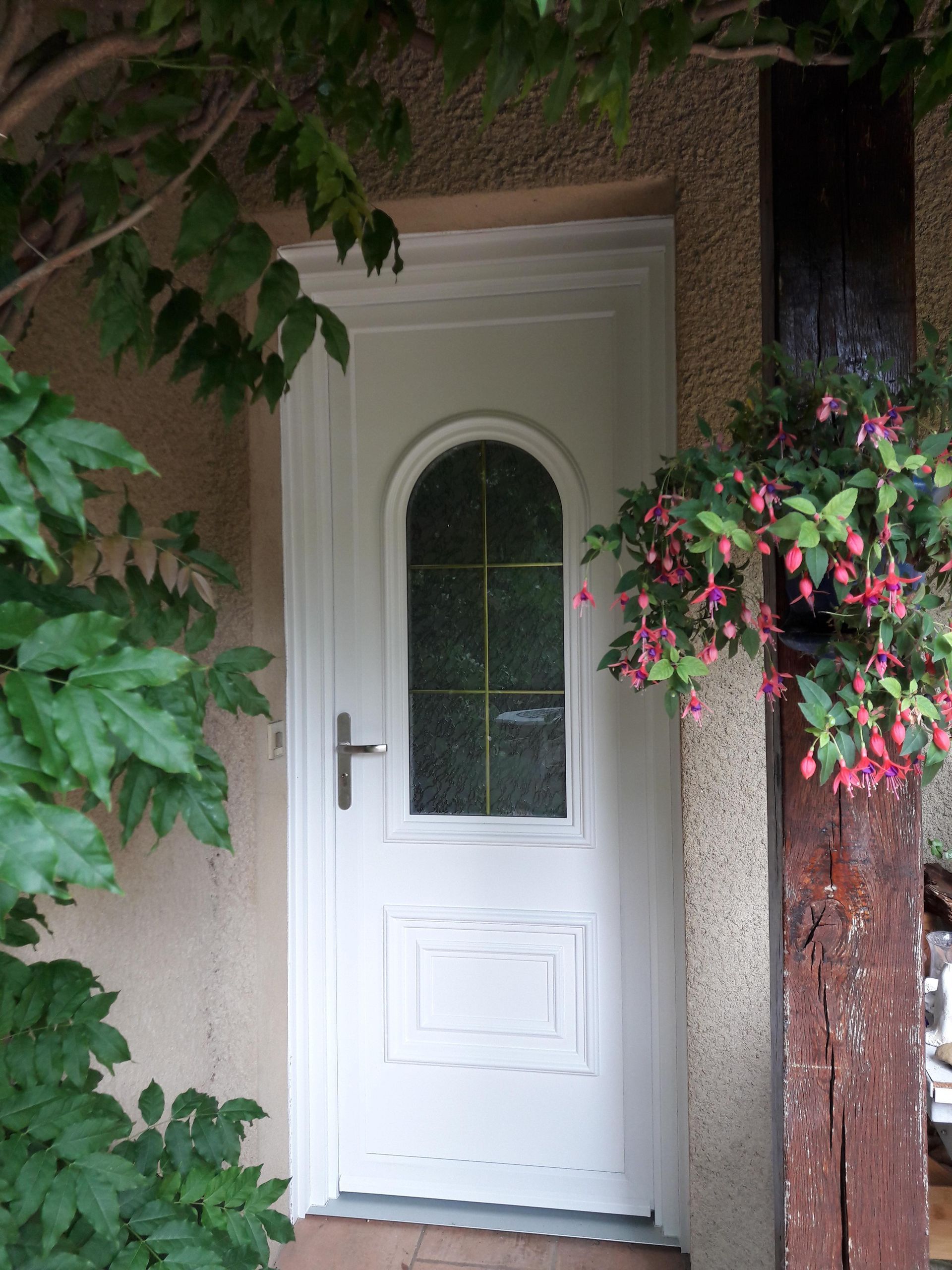 Une porte blanche avec une fenêtre cintrée, encadrée de feuillage vert et d'un fuchsia suspendu à un poteau en bois.