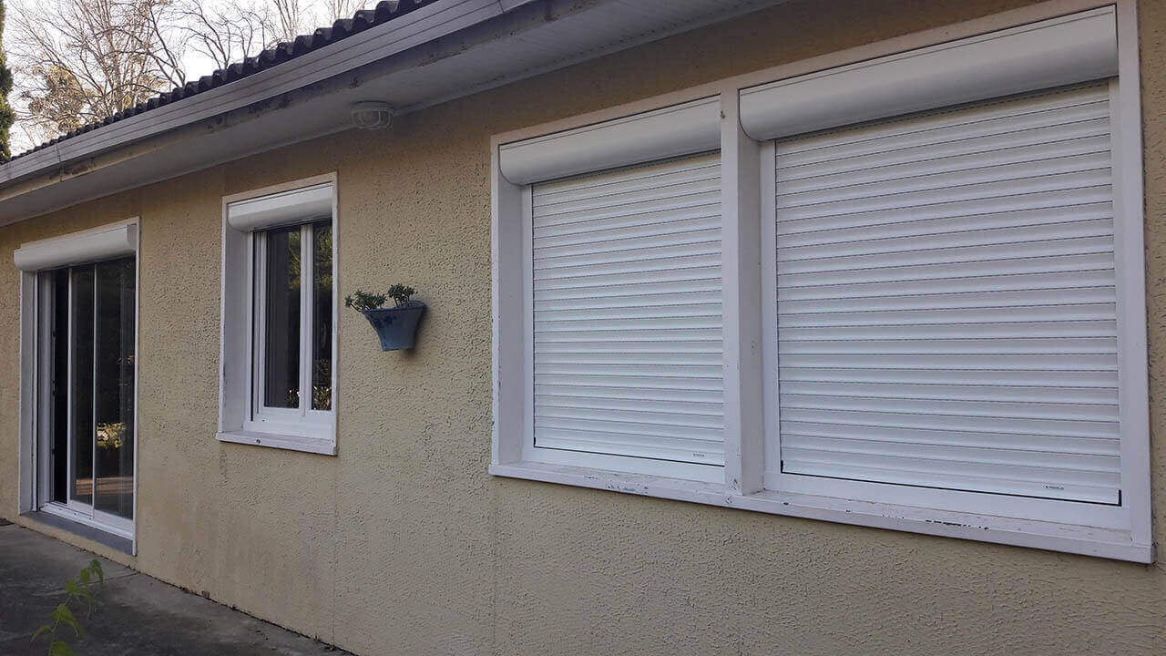Vue extérieure d'un mur de maison beige comportant trois fenêtres avec volets roulants blancs fermés et une petite jardinière murale.