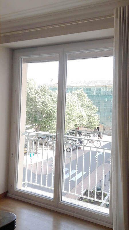 Une porte vitrée à cadre blanc s'ouvrant sur un balcon donnant sur une rue arborée avec des voitures garées.