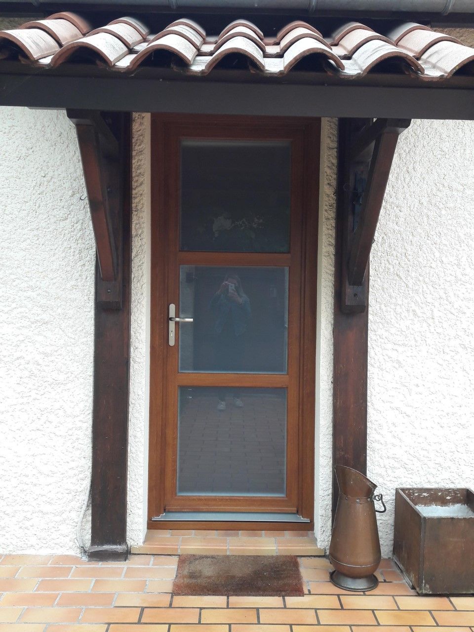 Une porte en bois brun avec une moustiquaire, sous un petit porche au toit de tuiles, contre un mur en stuc blanc avec un sol carrelé.
