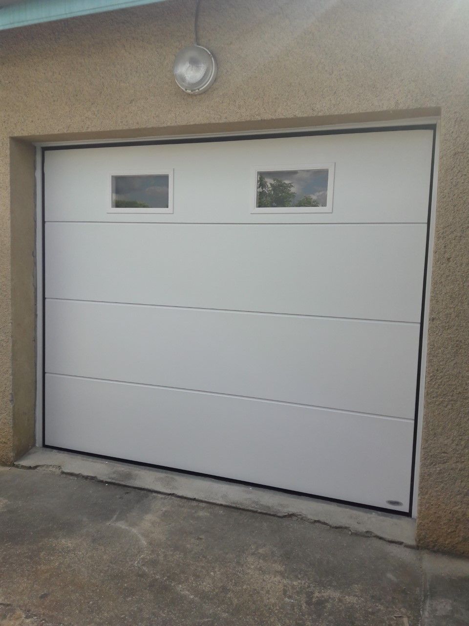Porte de garage sectionnelle blanche avec deux fenêtres rectangulaires, fixée sur un mur beige texturé au-dessus d'une allée en béton.
