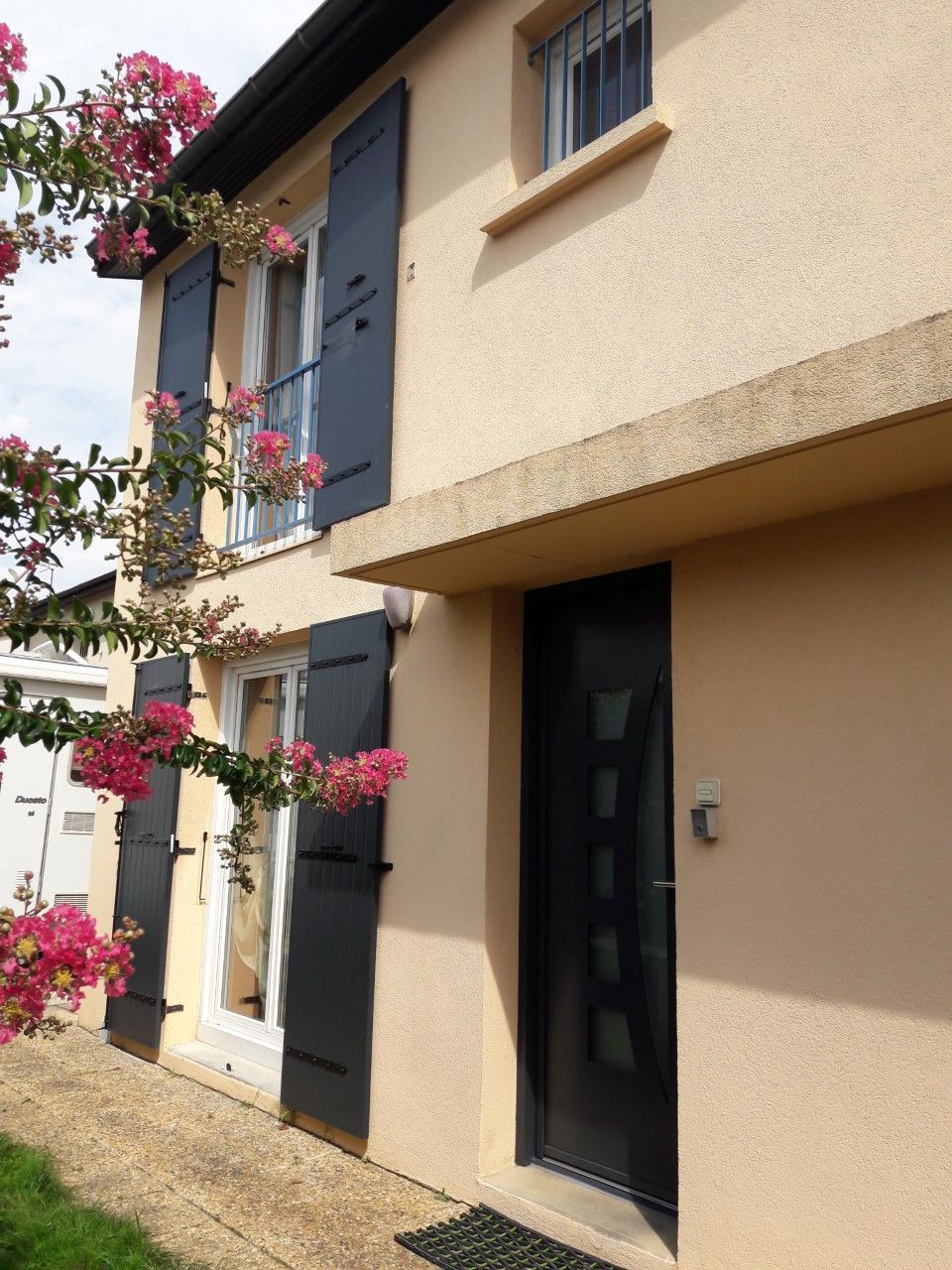 Une maison beige à deux étages avec des volets noirs, une porte d'entrée sombre et un lilas des Indes en fleurs au premier plan.