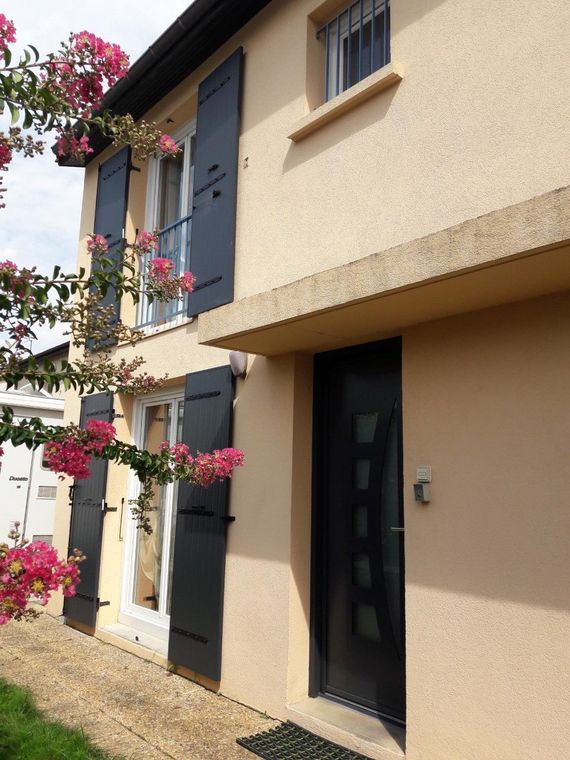 Une maison à deux étages en stuc beige avec des volets foncés, une porte d'entrée sombre et un lilas des Indes en fleurs au premier plan.