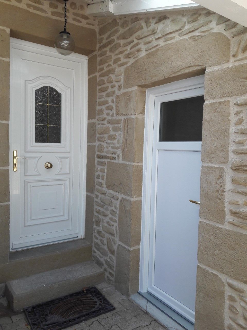 Deux portes blanches encastrées dans un mur de pierre, avec une petite marche et un paillasson devant la porte d'entrée principale.