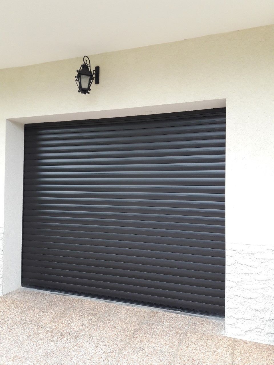 Une porte de garage noire à lames horizontales sur un bâtiment de couleur claire, avec une lampe extérieure fixée au mur au-dessus.