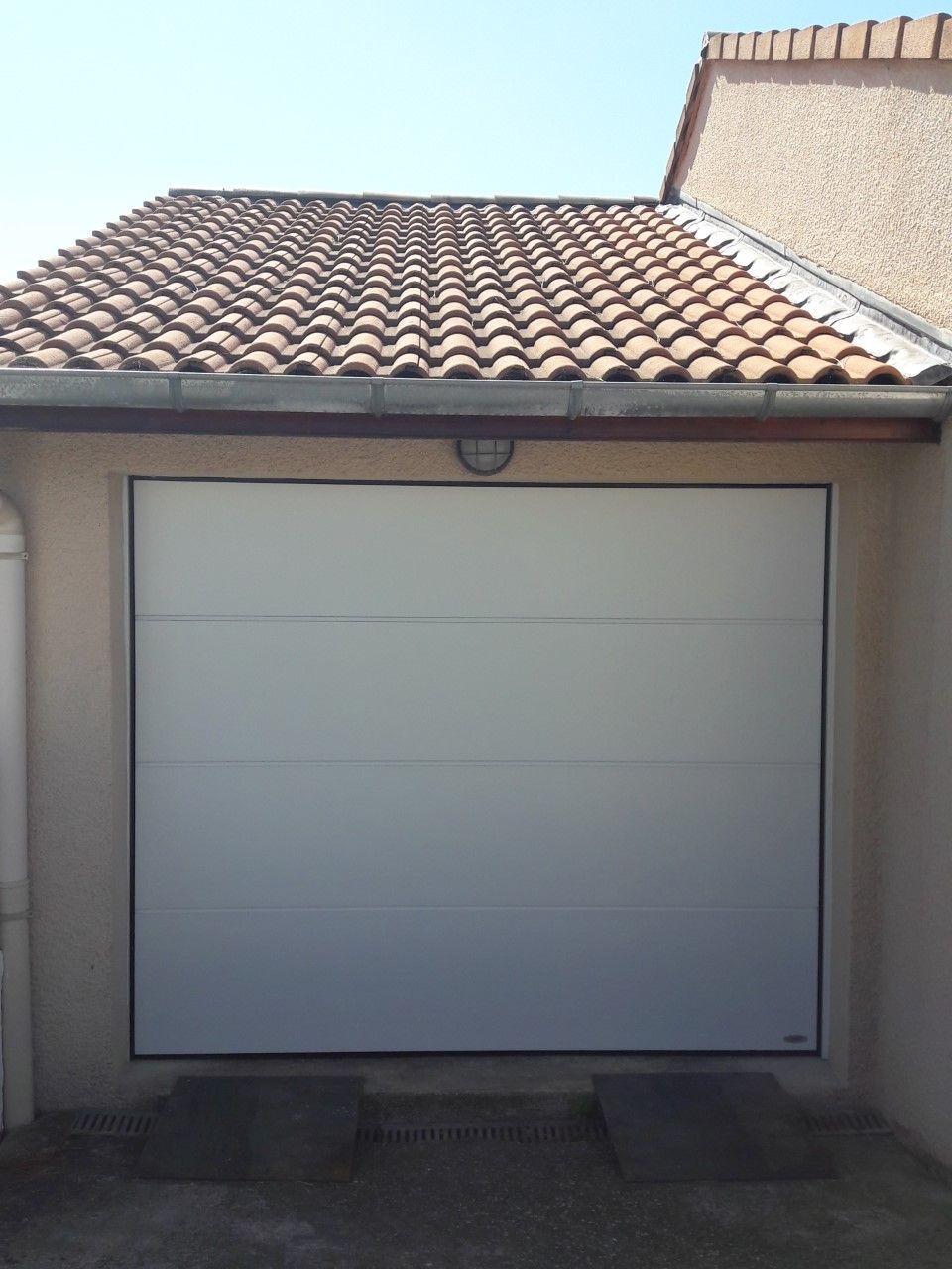 Une porte de garage blanche à panneaux horizontaux installée sur une maison avec un toit de tuiles et des murs en stuc clair.
