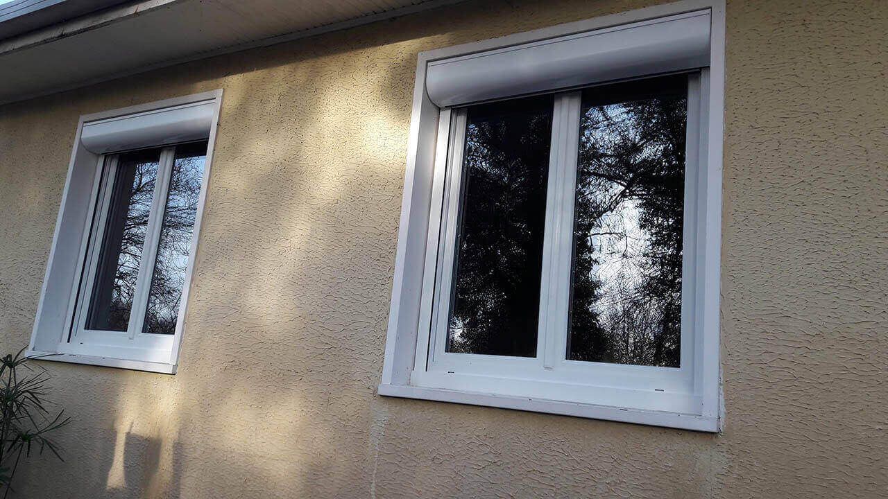 Deux fenêtres à cadre blanc avec volets roulants extérieurs installés sur un mur en stuc texturé de couleur claire.