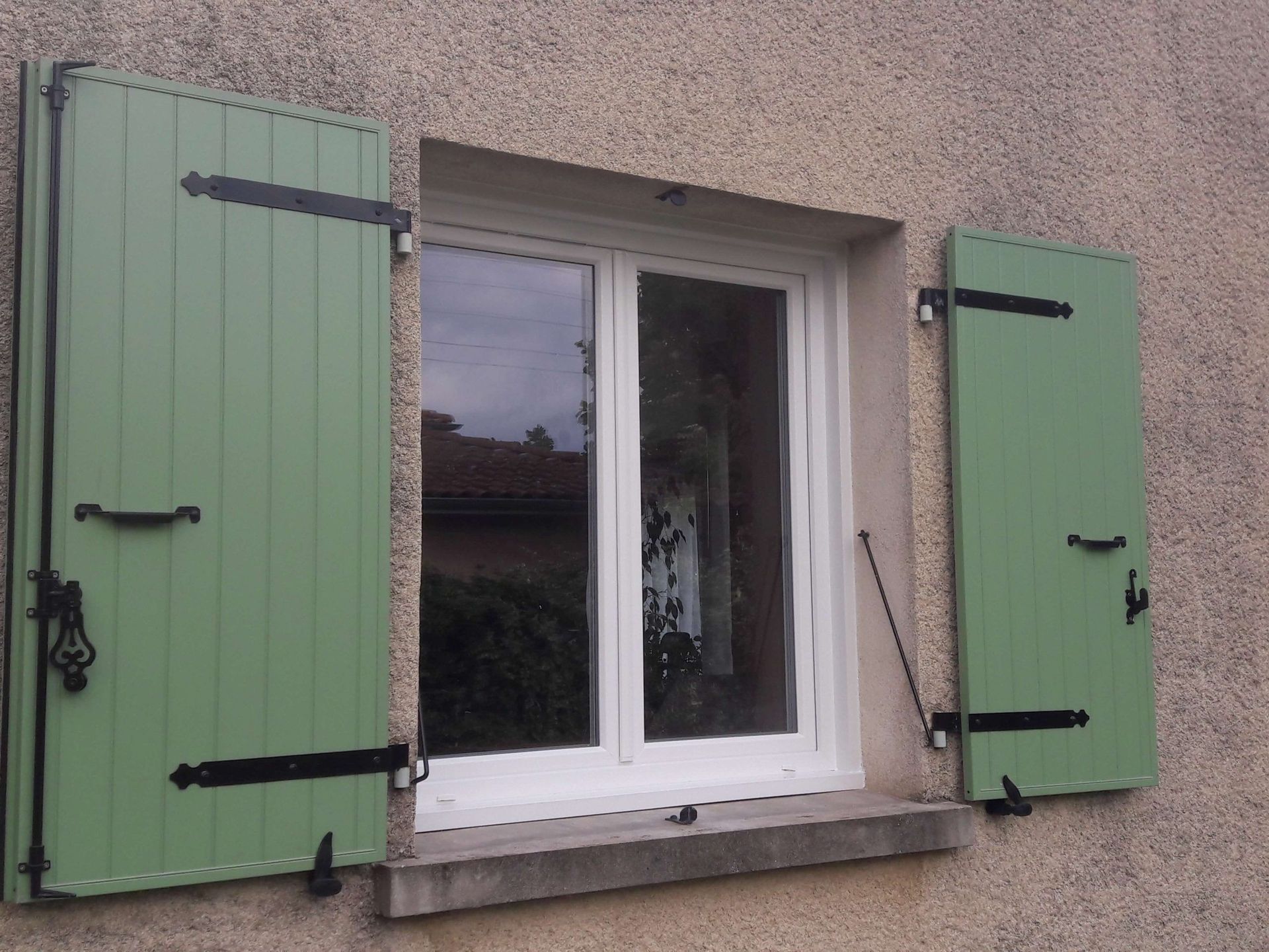Un cadre de fenêtre blanc est encastré dans un mur en stuc, flanqué de deux volets ouverts en bois vert sauge avec des ferrures en fer noir.