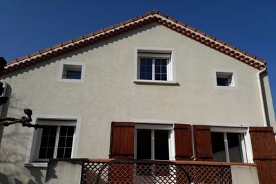 La façade d'une maison aux tons clairs, avec un toit pointu, cinq fenêtres et des volets bruns, se détache sur un ciel bleu dégagé.