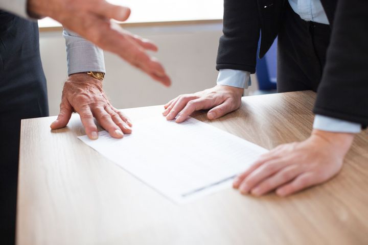 Dos personas examinan un documento sobre una mesa de madera. Una hace un gesto con la mano, la otra apoya las manos