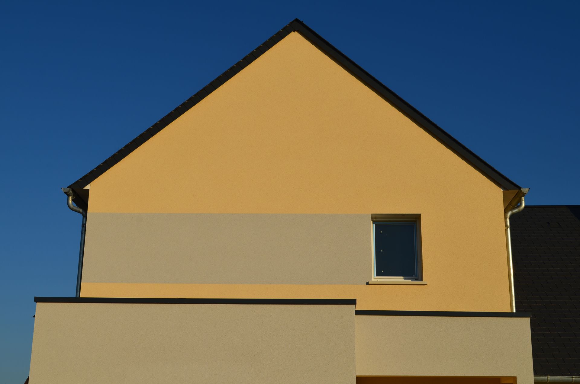 Façade de maison jaune avec un bord de toit sombre et une petite fenêtre.