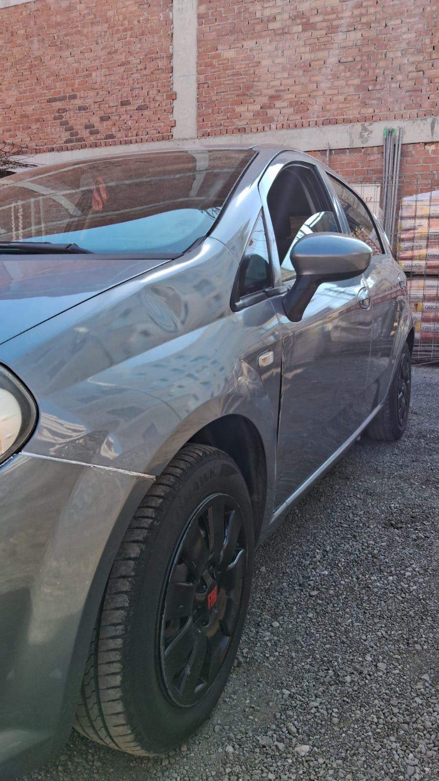 Coche Fiat gris aparcado sobre grava, llantas negras, cerca de una pared de ladrillos.