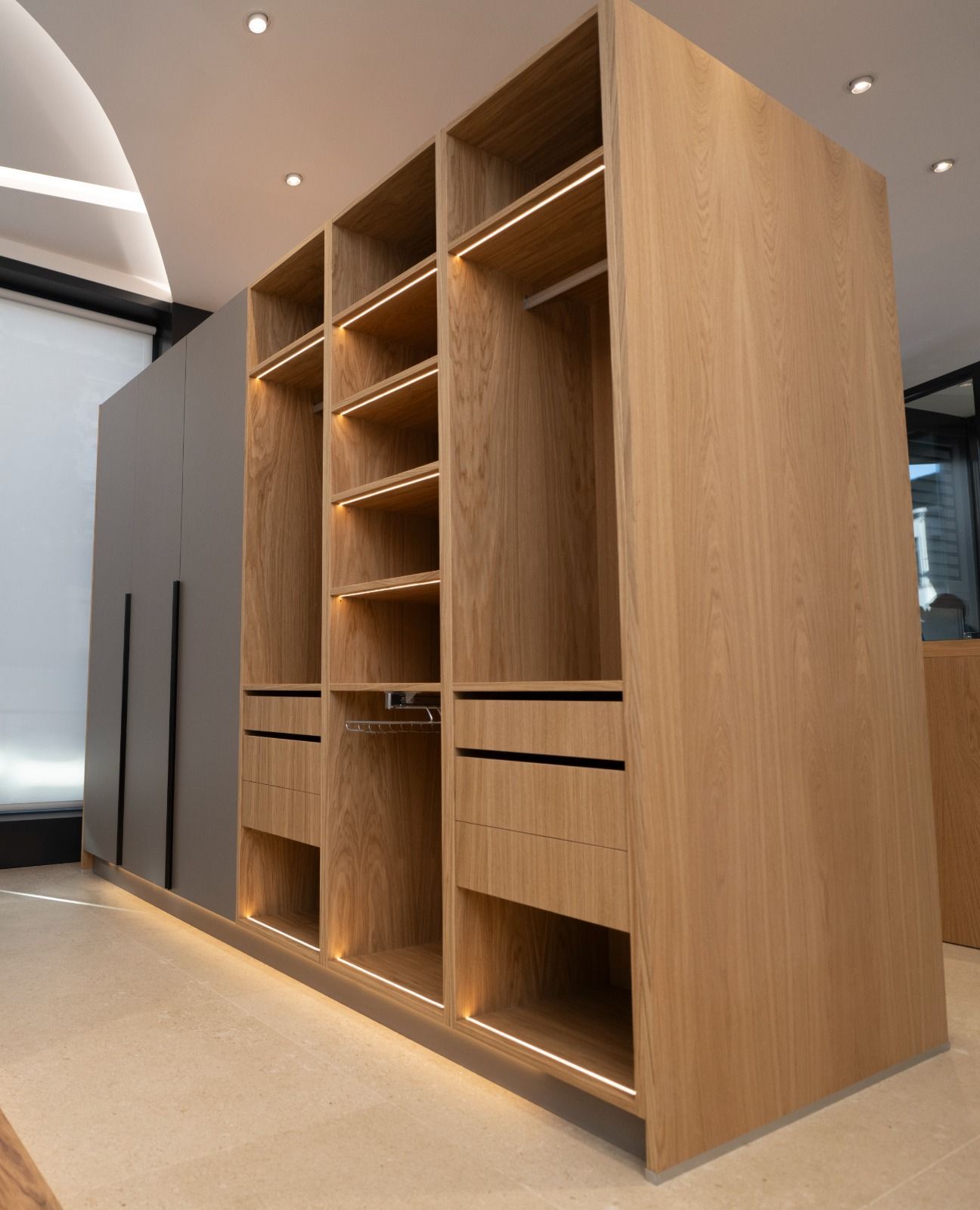 Armario de madera con estantes abiertos, cajones y espacio para colgar, iluminado con tiras de luz. Puertas grises a la izquierda.