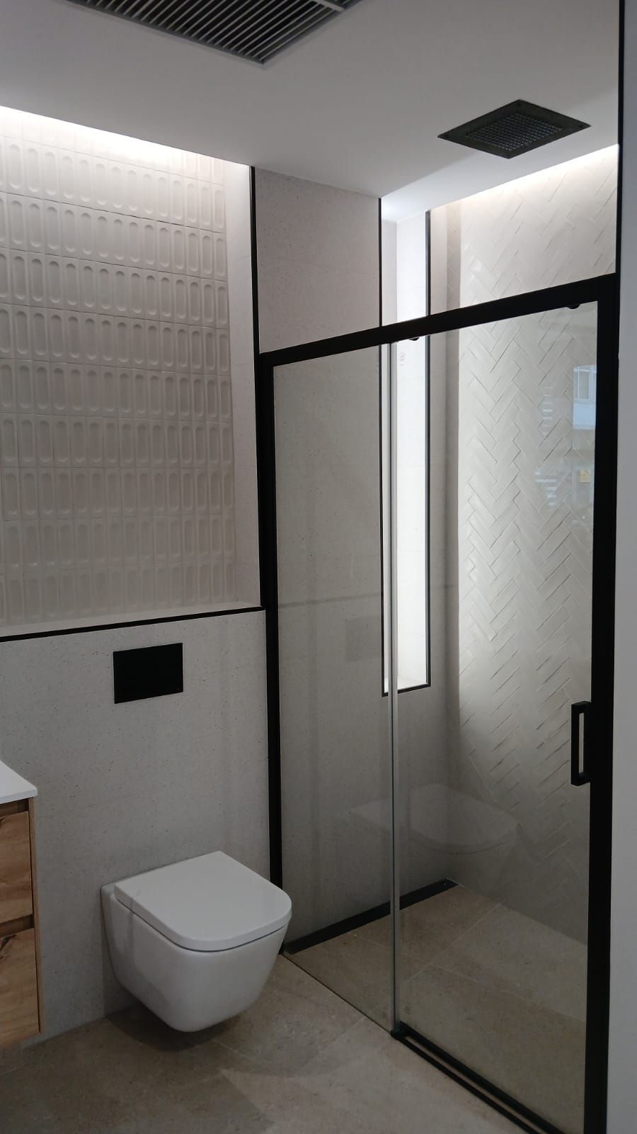 Baño moderno con ducha de cristal, marco negro, inodoro blanco, azulejos gris claro y tocador de madera.