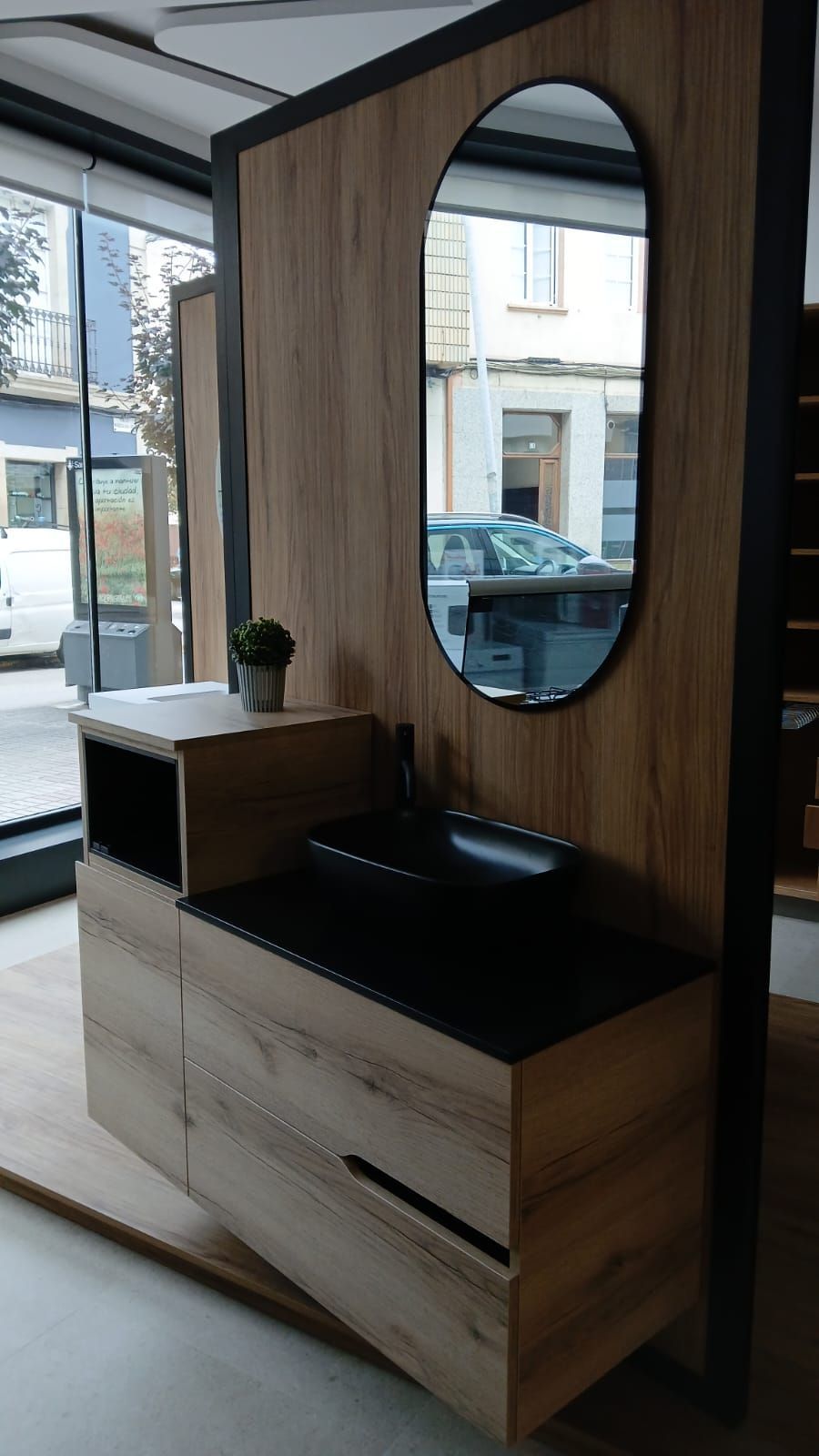 Mueble de baño con gabinetes de veta de madera, encimera negra, espejo ovalado.
