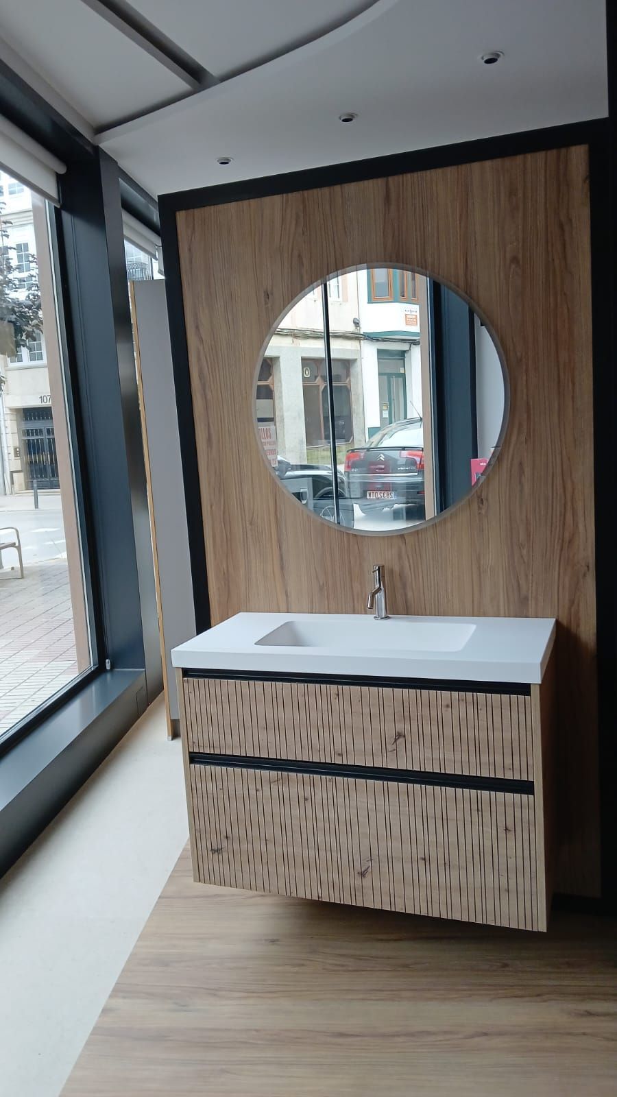 Mueble de baño con mueble de madera texturizada, espejo redondo y ventana con vista.