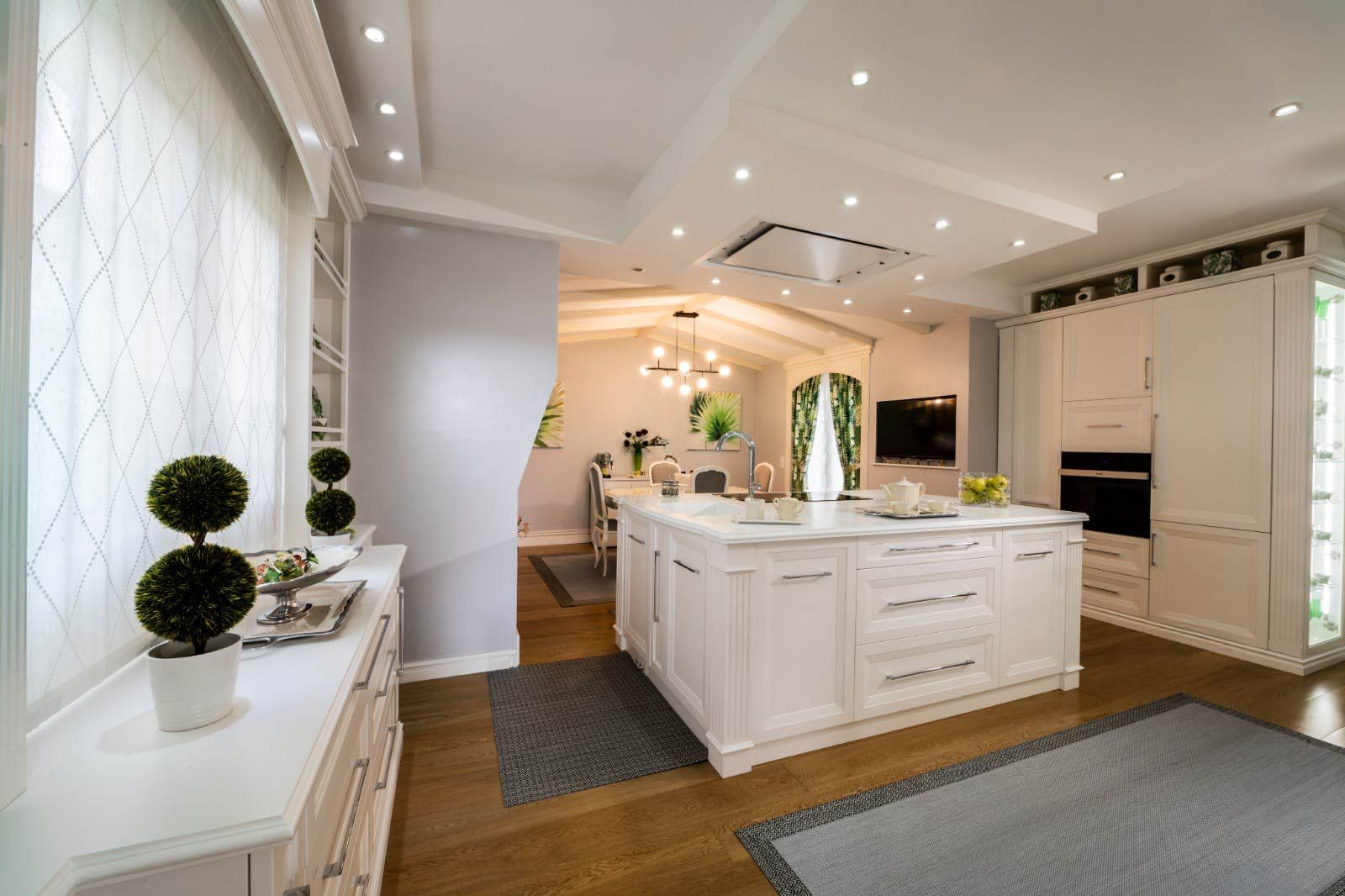 Cocina blanca luminosa y moderna con isla y gabinetes, pisos de madera, artefactos de iluminación y vista a la sala de estar.