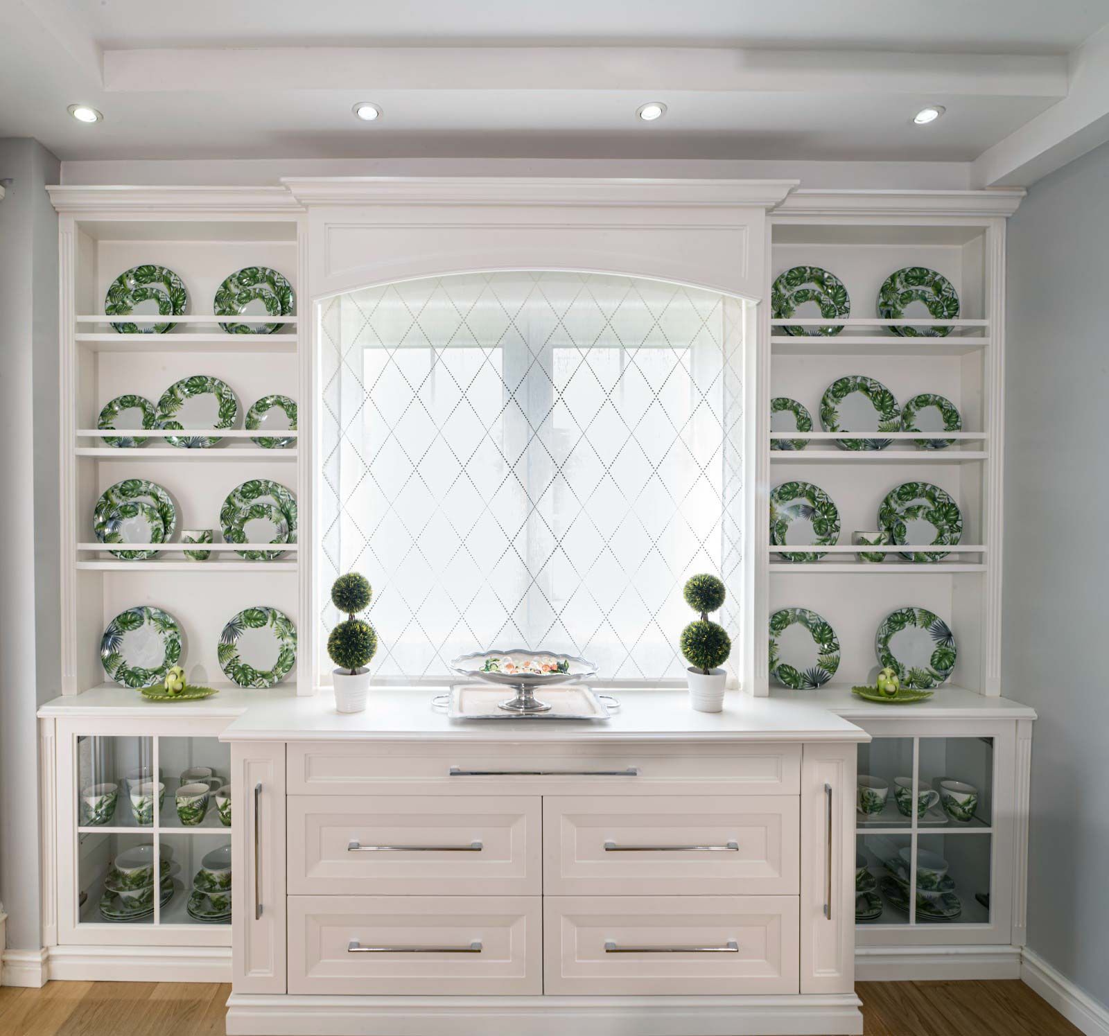 Aparador blanco con estantes para exposición de platos, ventana con diseño de rombos y mostrador con cajones y dos topiarios.