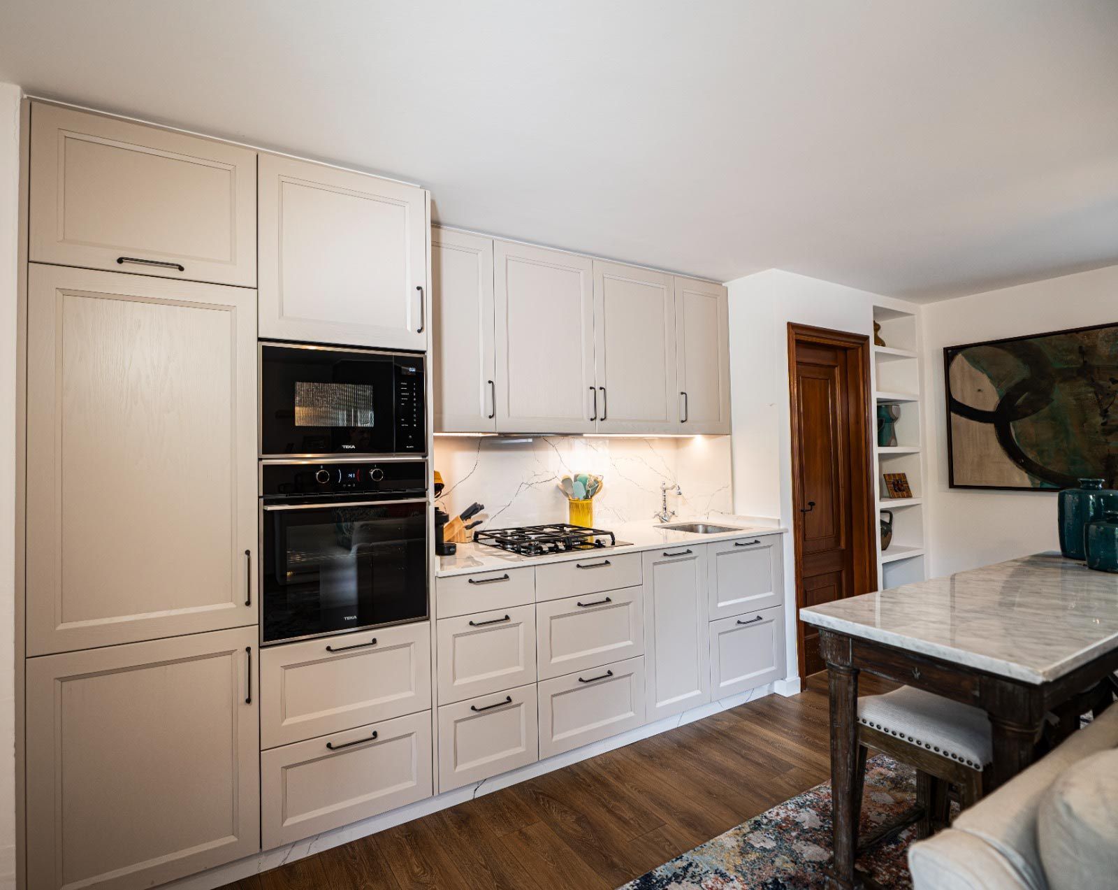 Cocina beige con horno, gabinetes y mesa de comedor con cubierta de mármol sobre piso de madera.