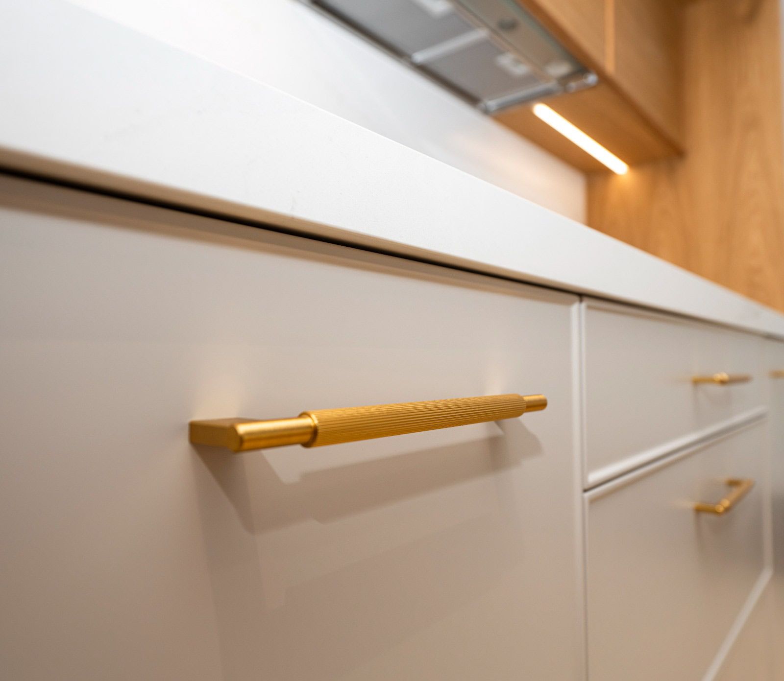 Primer plano de gabinetes de cocina blancos con herrajes dorados. Encimera y estante de madera al fondo.