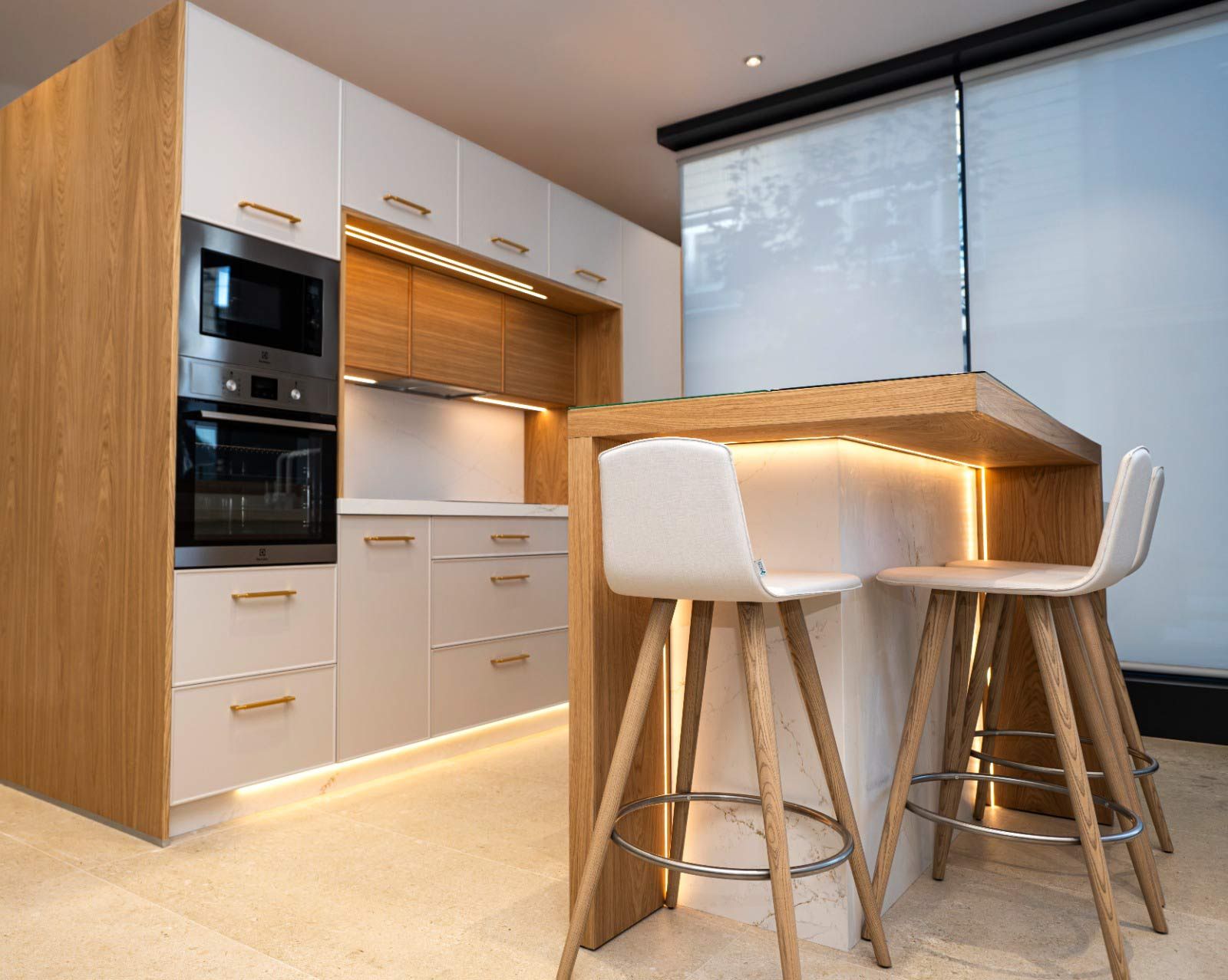 Cocina moderna con gabinetes blancos, detalles de madera, isla con taburetes y electrodomésticos empotrados.