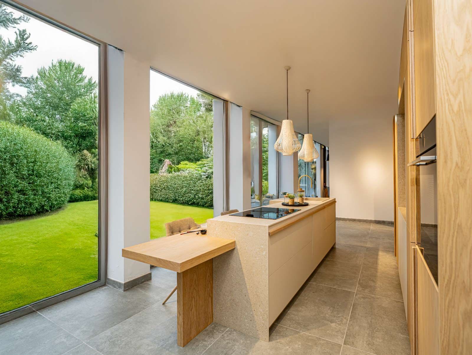 Cocina moderna con amplios ventanales con vistas a un exuberante jardín. Gabinetes de madera clara, isla con estufa y una pequeña mesa de comedor.