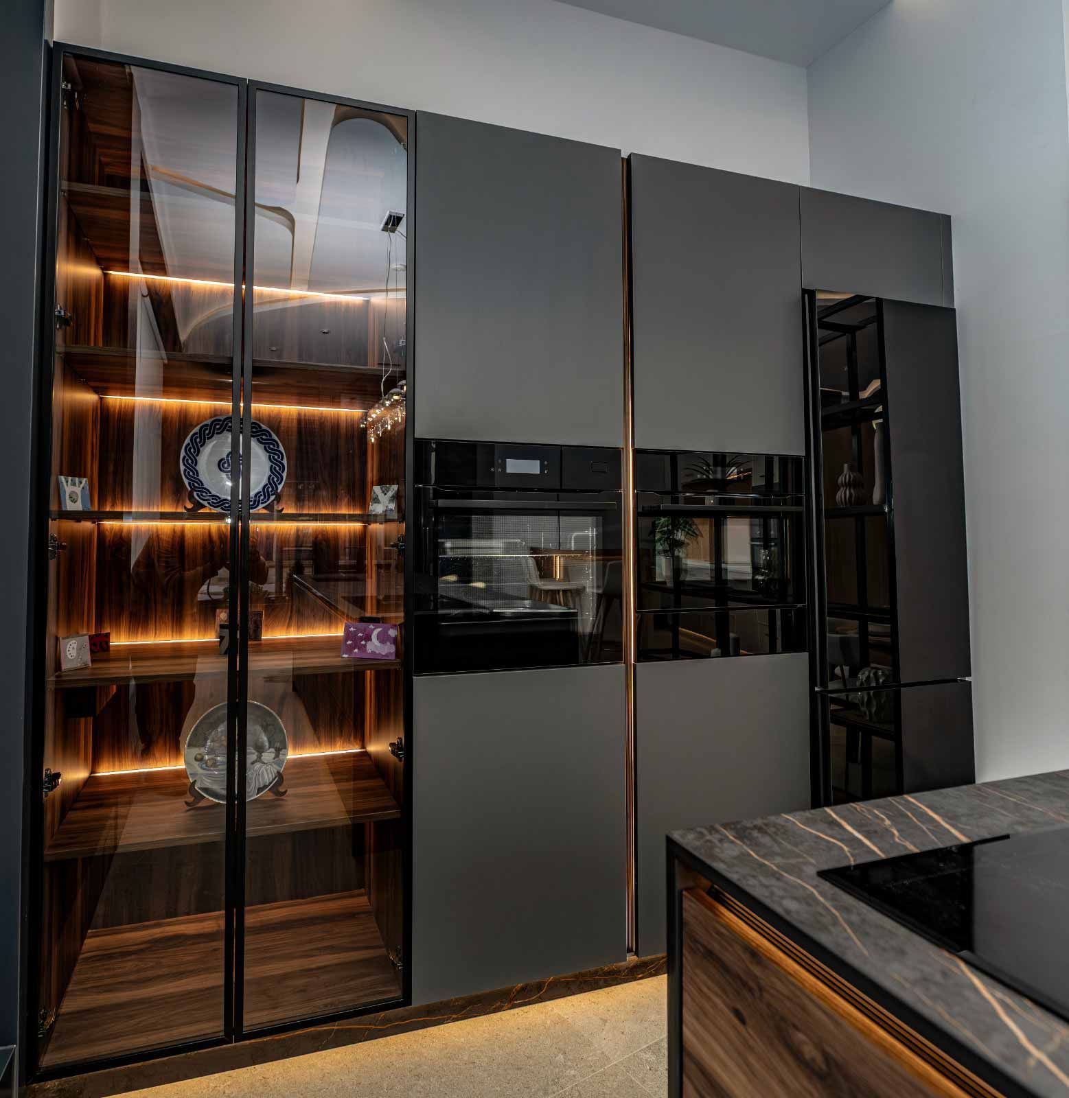 Cocina moderna con gabinetes gris oscuro, vitrina y hornos empotrados. Detalles dorados e iluminación cálida.