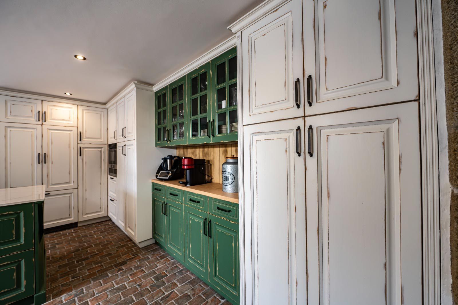 Cocina con gabinetes blancos desgastados y gabinetes con detalles verdes, piso de ladrillo.