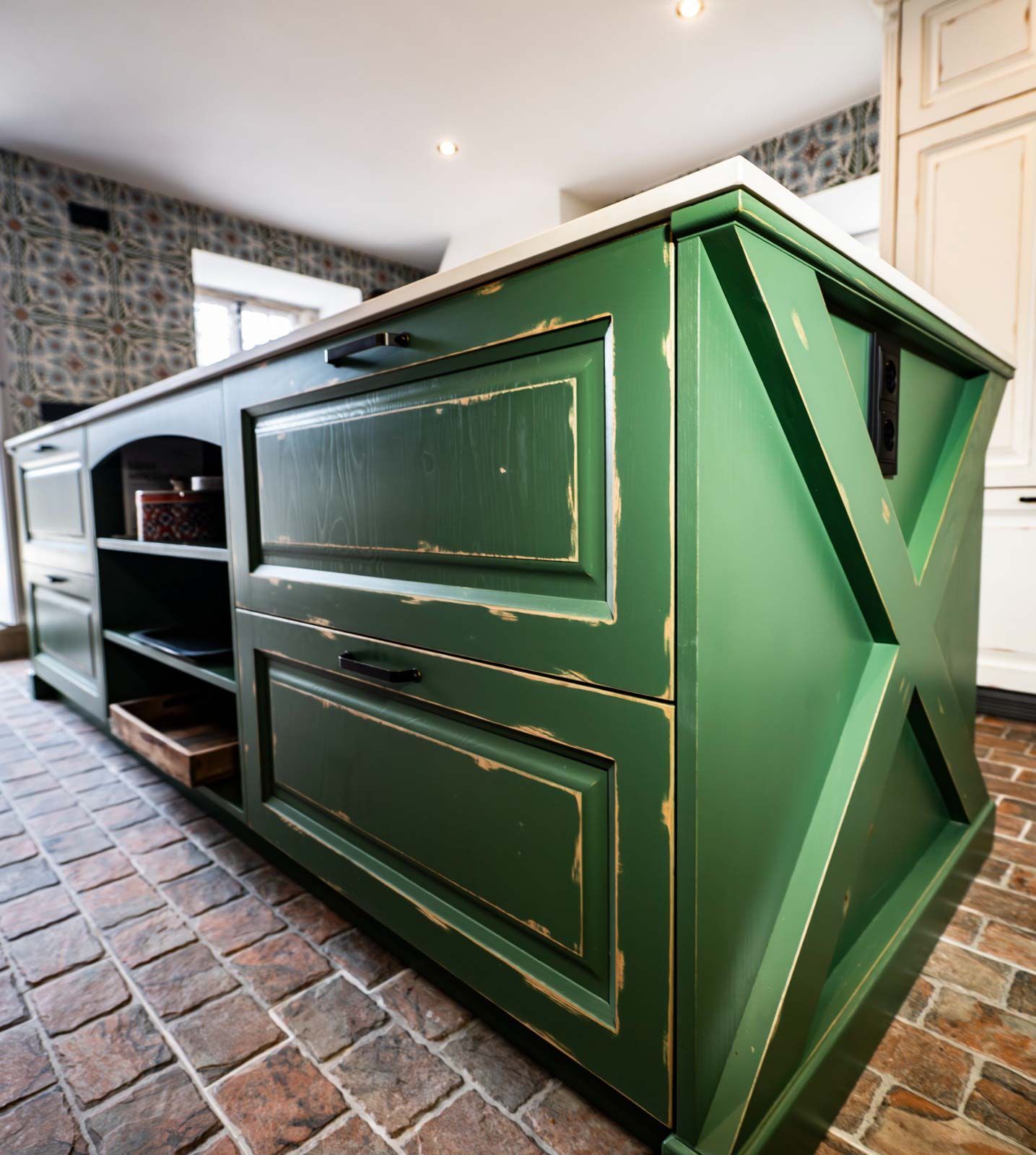 Isla de cocina verde con acabado envejecido, herrajes negros y panel lateral en forma de X, sobre piso de ladrillo.
