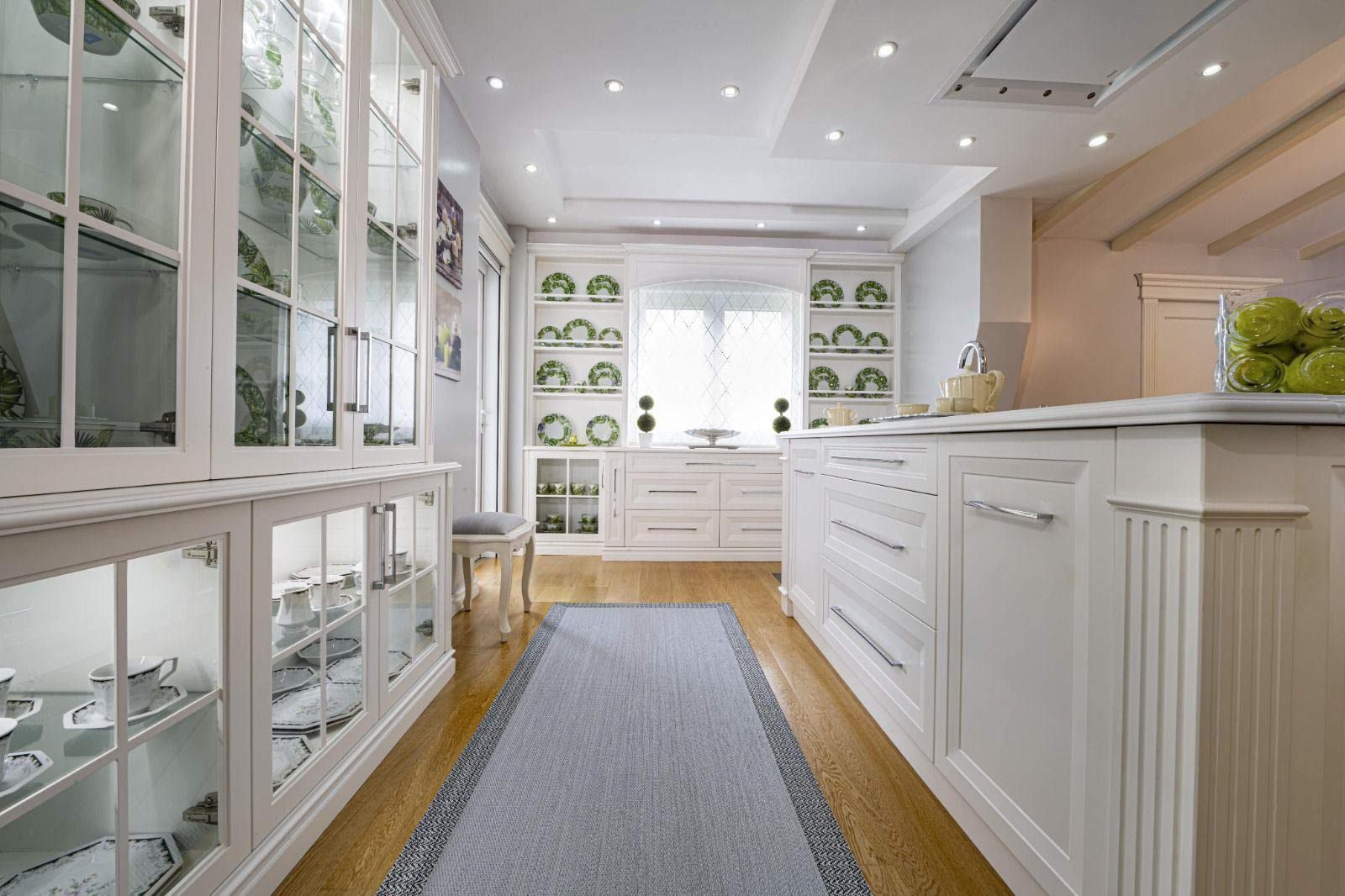 Cocina blanca con gabinetes de vidrio que exhiben vajilla, pisos de madera y una alfombra azul.