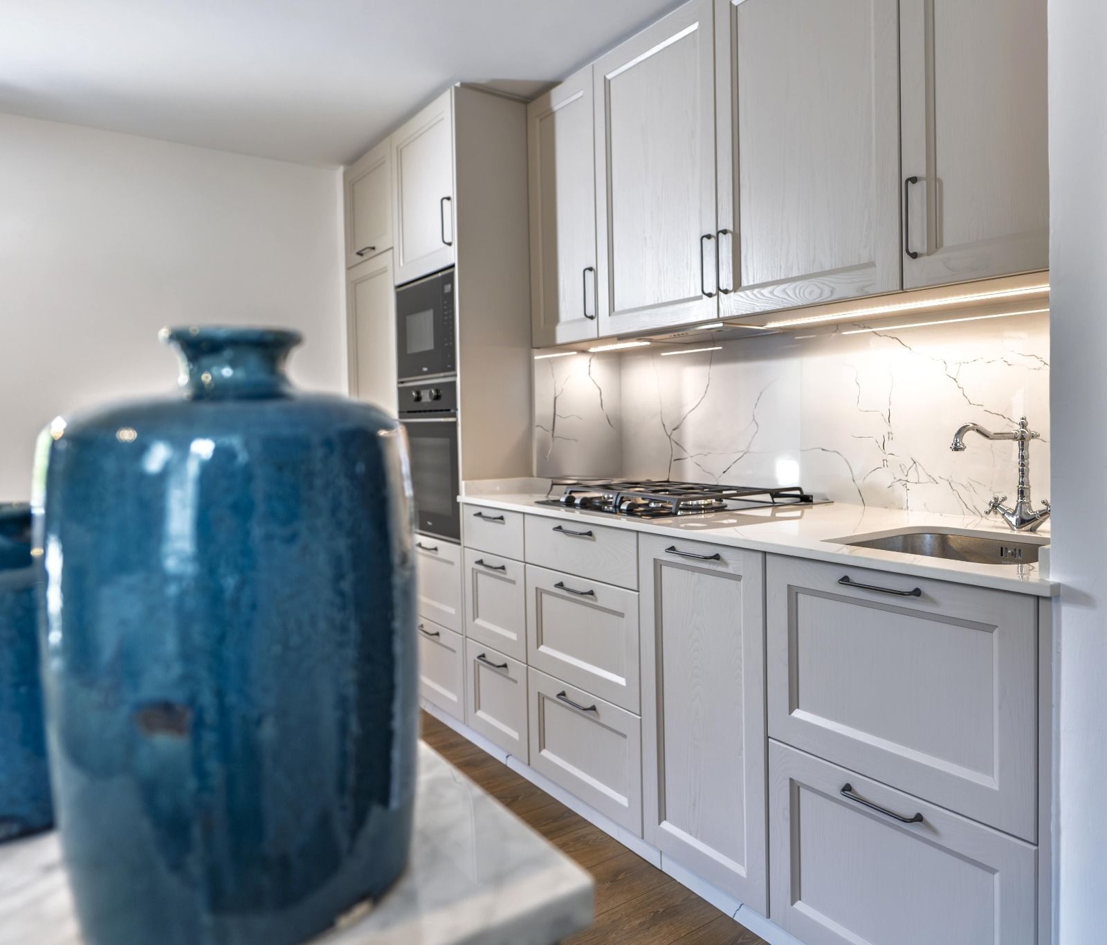 Cocina de color claro con muebles, encimera de mármol, horno empotrado y jarrón azul en primer plano.