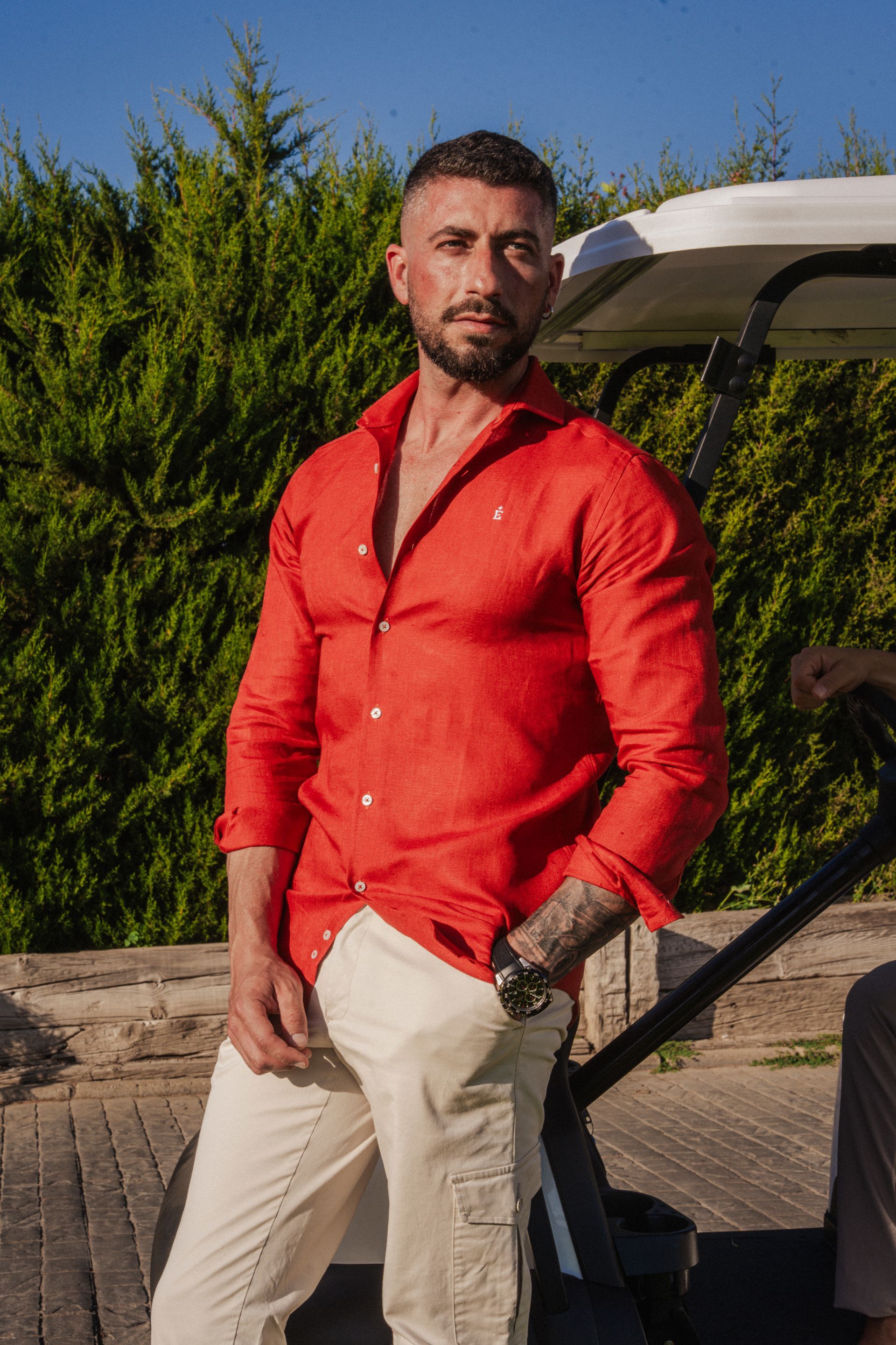 Un hombre está de pie al aire libre al atardecer, con una camisa rosa, pantalones grises y gafas de sol, cerca de unas palmeras.