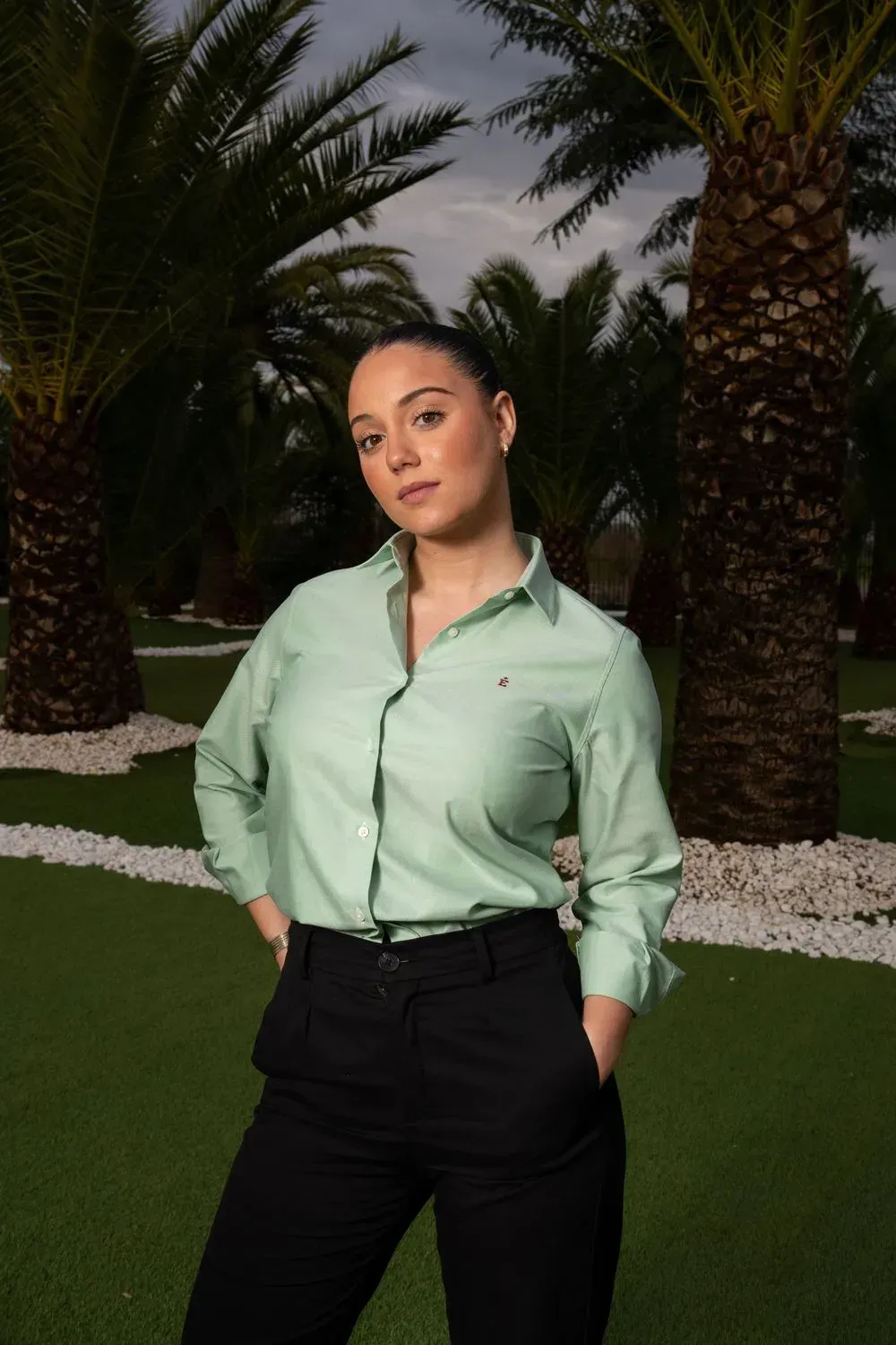 Una mujer con una camisa verde y pantalones negros está parada frente a unas palmeras.