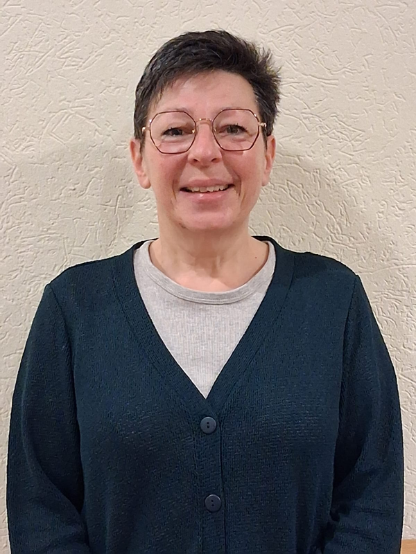 Frau mit Brille, lächelnd, mit Halskette und gemustertem Oberteil, in heller Umgebung.