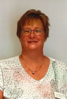 Frau mit Brille, lächelnd, mit Halskette und gemustertem Oberteil, in heller Umgebung.