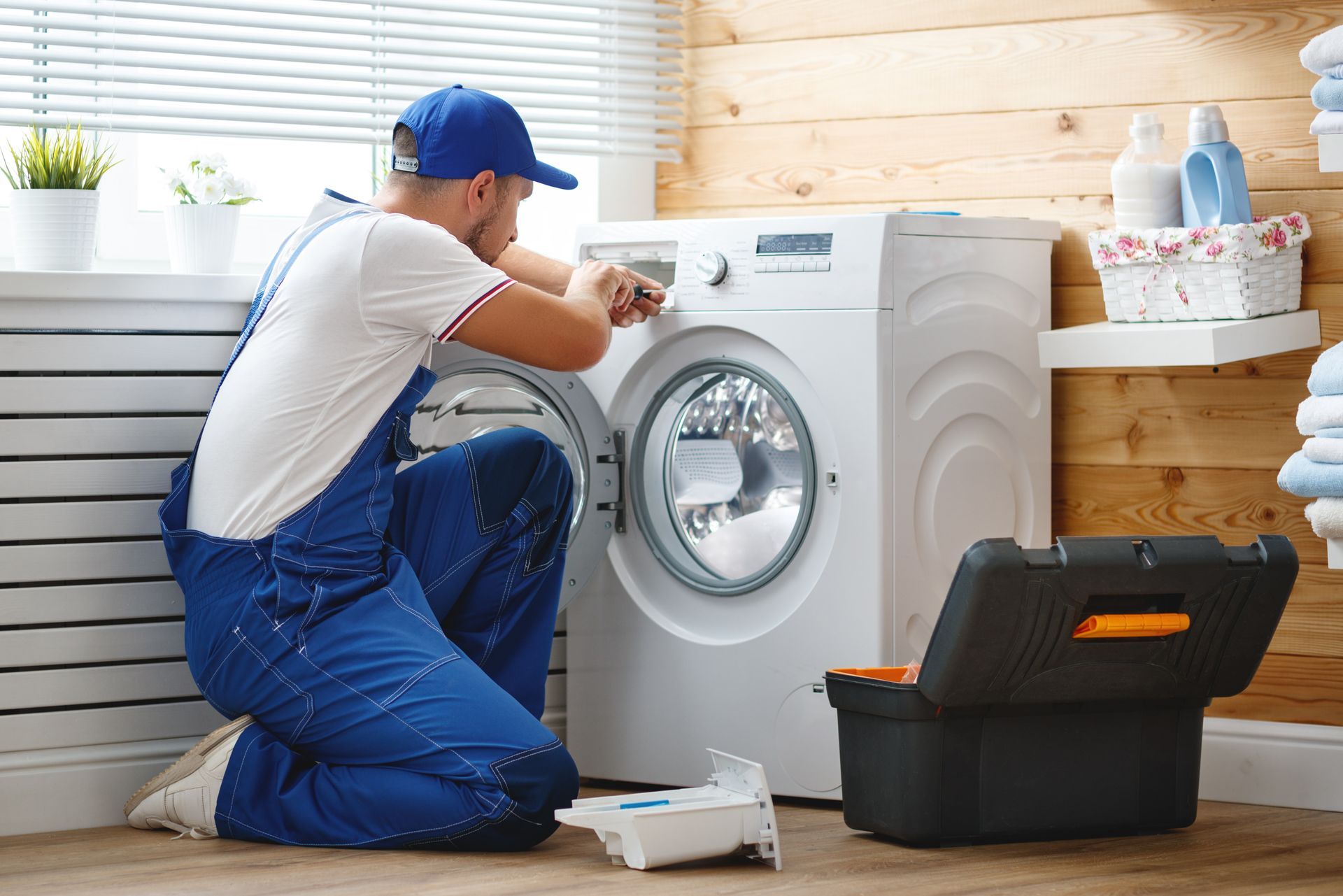 Un homme en salopette bleue répare une machine à laver blanche, à genoux avec une boîte à outils devant elle.