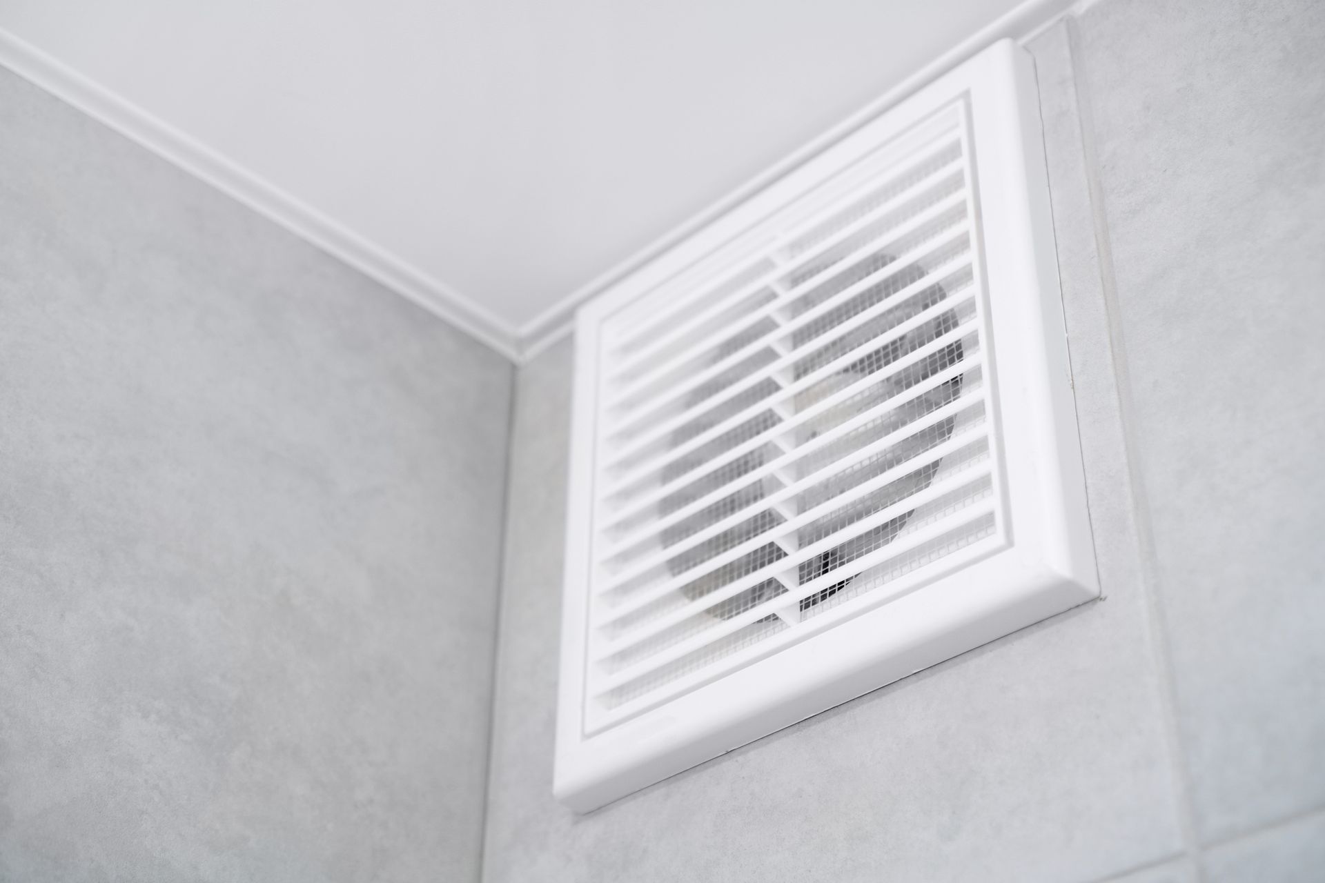 Grille de ventilation de salle de bains blanche sur un mur carrelé gris, près du plafond.