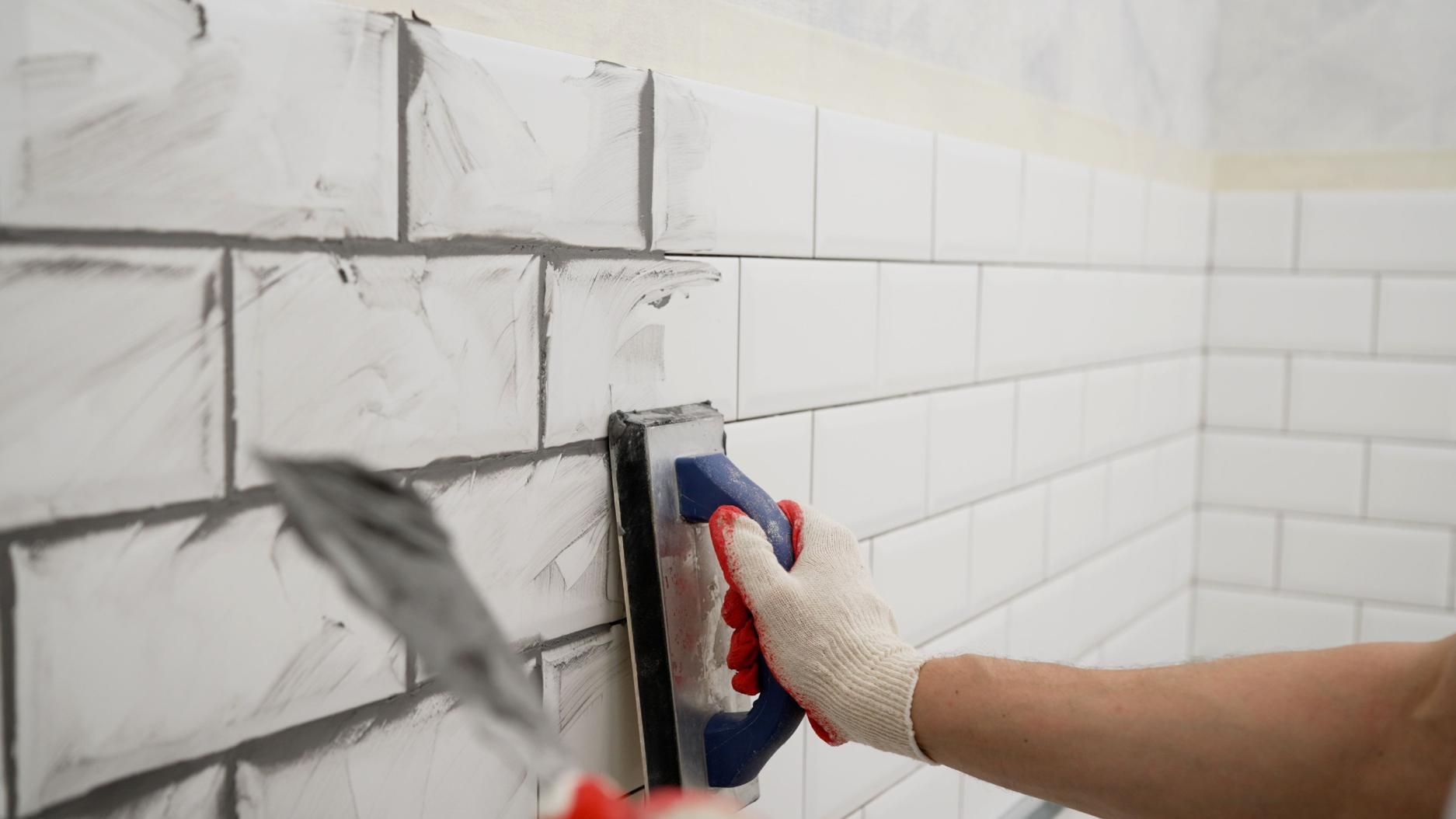 Persona aplicando mortero gris sobre una pared de azulejos blancos durante la instalación.