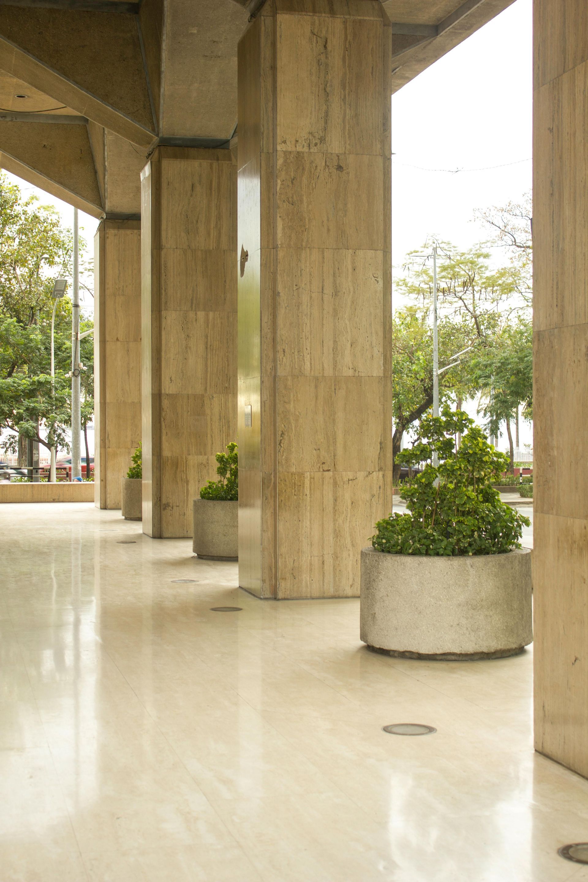 Pasarela cubierta con columnas de hormigón, suelo brillante y jardineras redondas junto a los árboles.