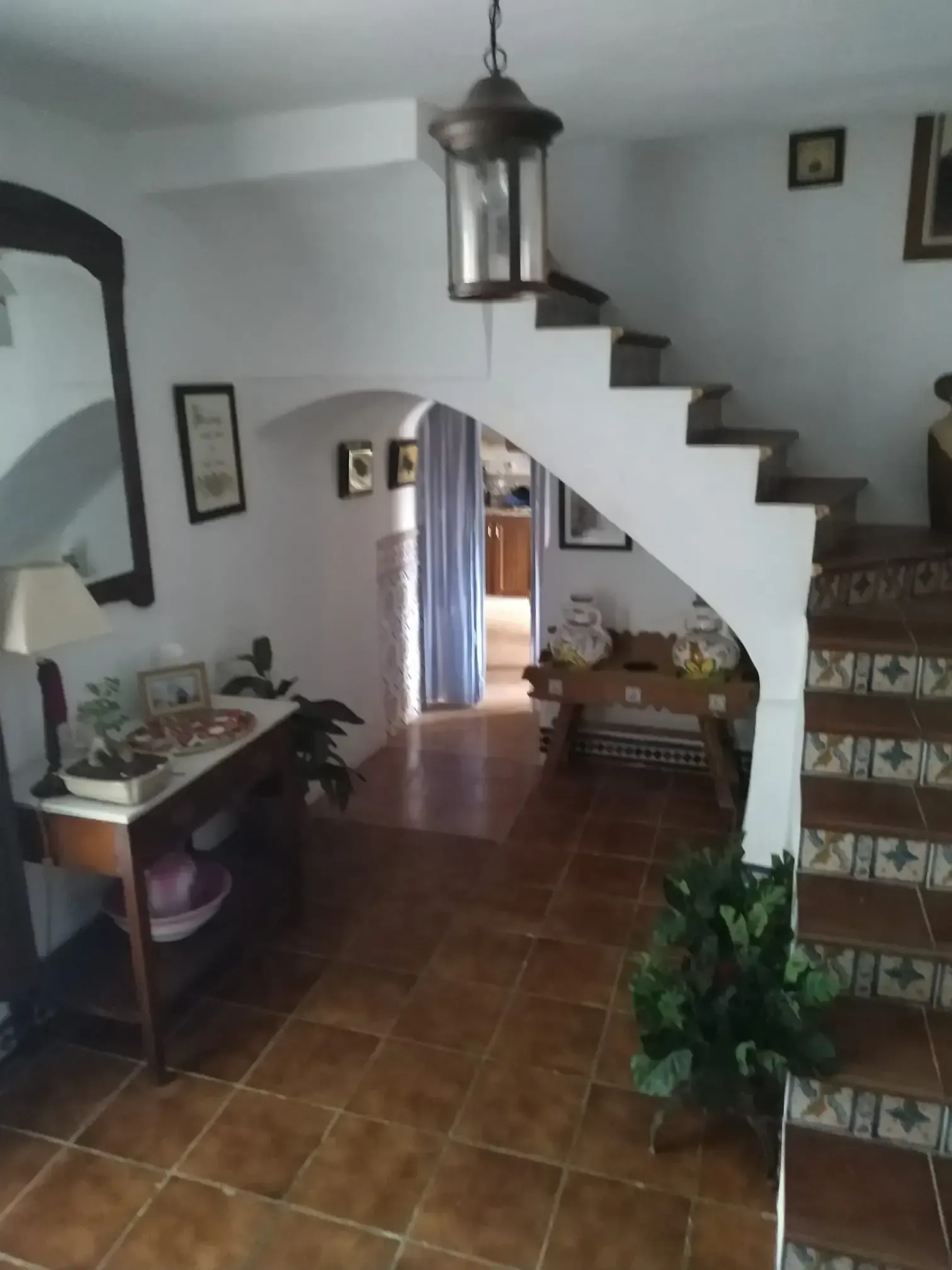 Vista interior de un pasillo con escalera, suelo de baldosas, arcos y una linterna colgante.