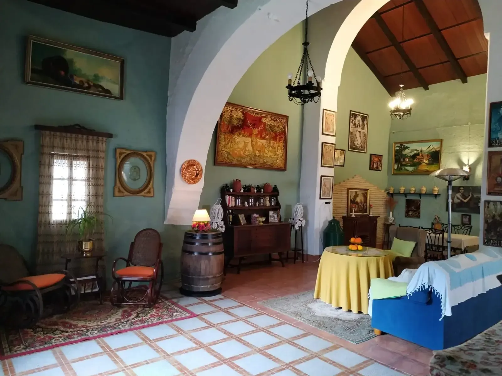 Habitación espaciosa con puertas arqueadas, cuadros, muebles antiguos y un barril. Paredes azul claro y suelo de baldosas.