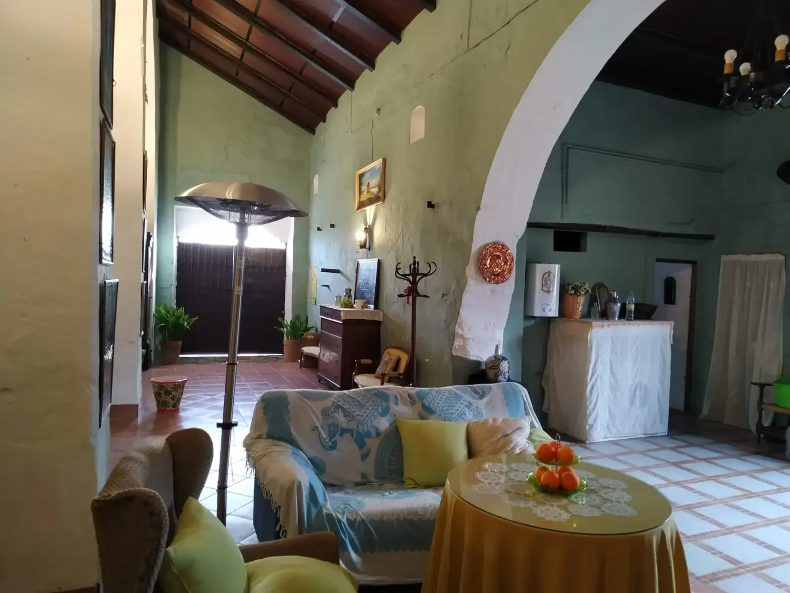 Habitación interior con arco, sofá, mesa y calefactor alto. Paredes verdes, suelo de baldosas, techo de madera.