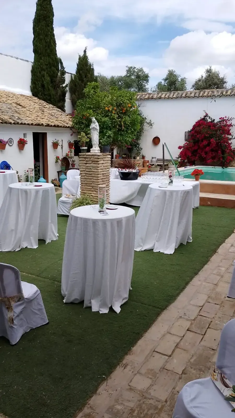Montaje de evento al aire libre con mesas cubiertas con manteles blancos sobre césped verde, una estatua y una piscina.