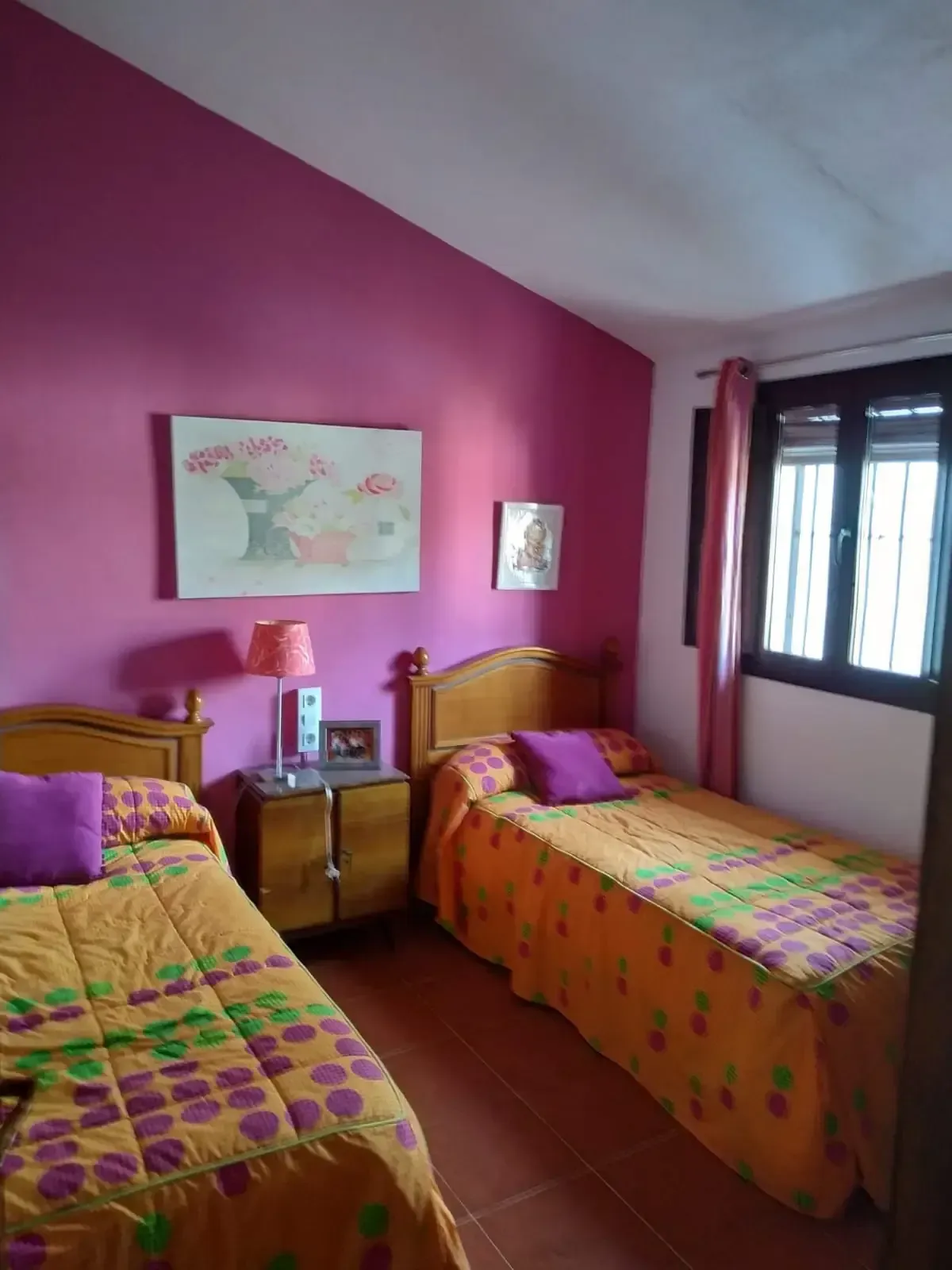 Dos camas individuales con edredones naranjas en una habitación con paredes magenta y una ventana.