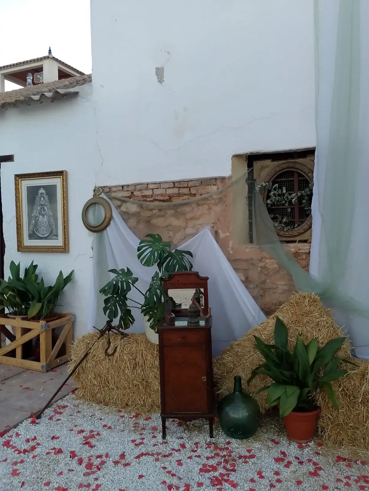 Escena rústica al aire libre con plantas, fardos de heno y muebles antiguos contra una pared blanca con un cuadro enmarcado.